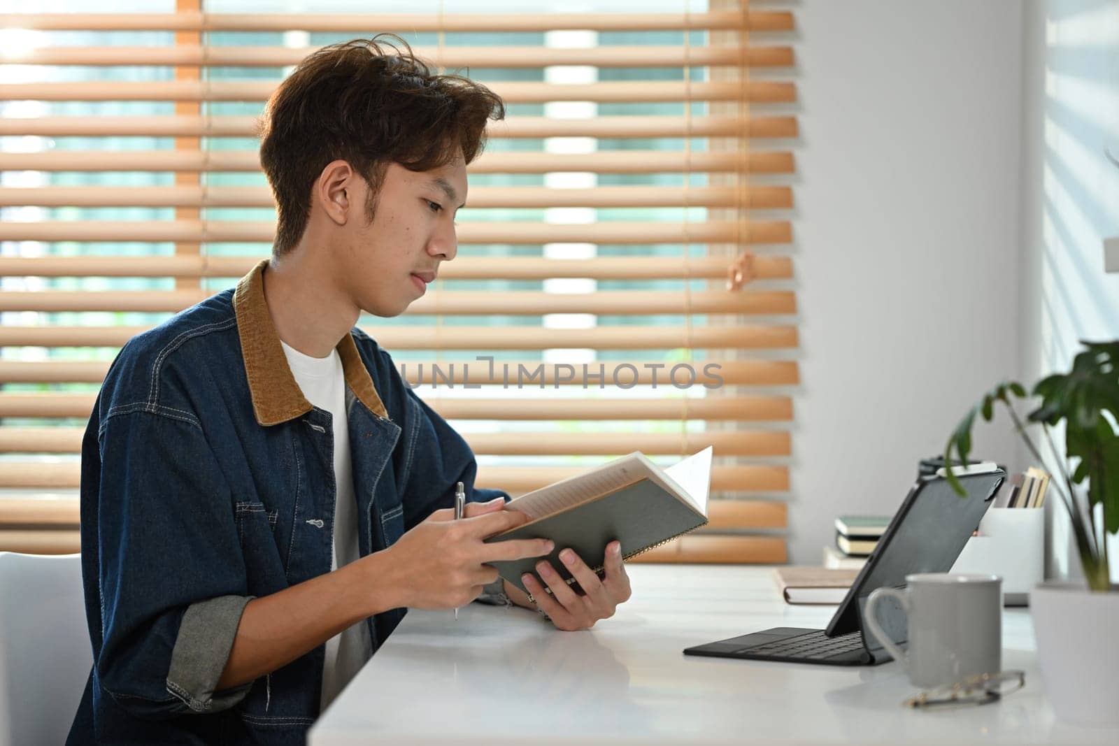 Asian male university student doing homework sitting at table with laptop computer next to window at home by prathanchorruangsak