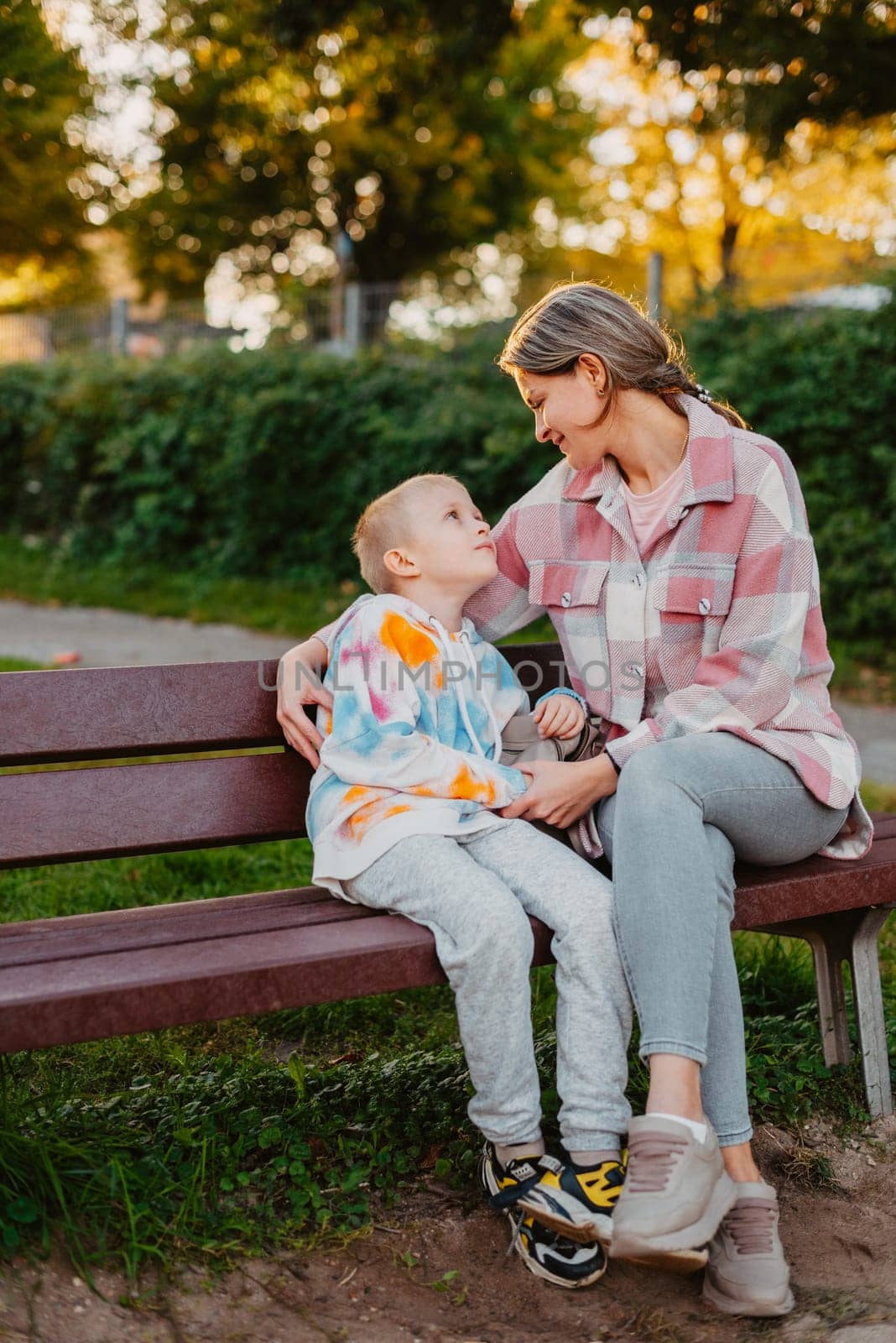 mother and son sit on a park bench in the rays of the setting sun. the concept of a family. Mother's Day. beautiful girl (mother) with a boy (son) in the park in the park are sitting on a bench at sunset.