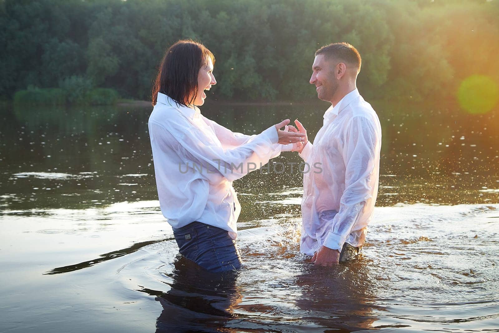 Beautiful adult couple has fun in nature in the water in a river or lake in the summer evening at sunset. A guy and a girl swim and relax outdoors in clothes in white shirts and jeans