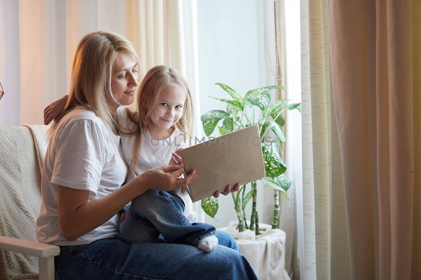 Happy loving family with mother and daughter reading book in living room. Woman mom and small child girl having convercation and studing inside of home