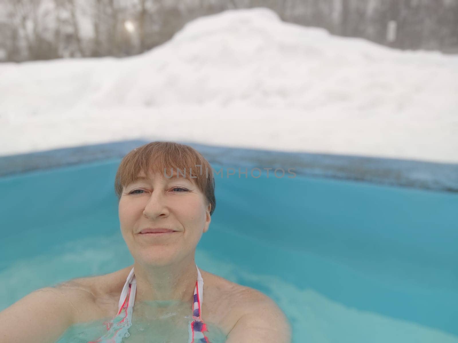 Adult mature woman takes selfie in a pool with warm hot termal mineral water in winter and white snow around. Wellness center and the concept of health care. Travel, recreation, medicine and rest