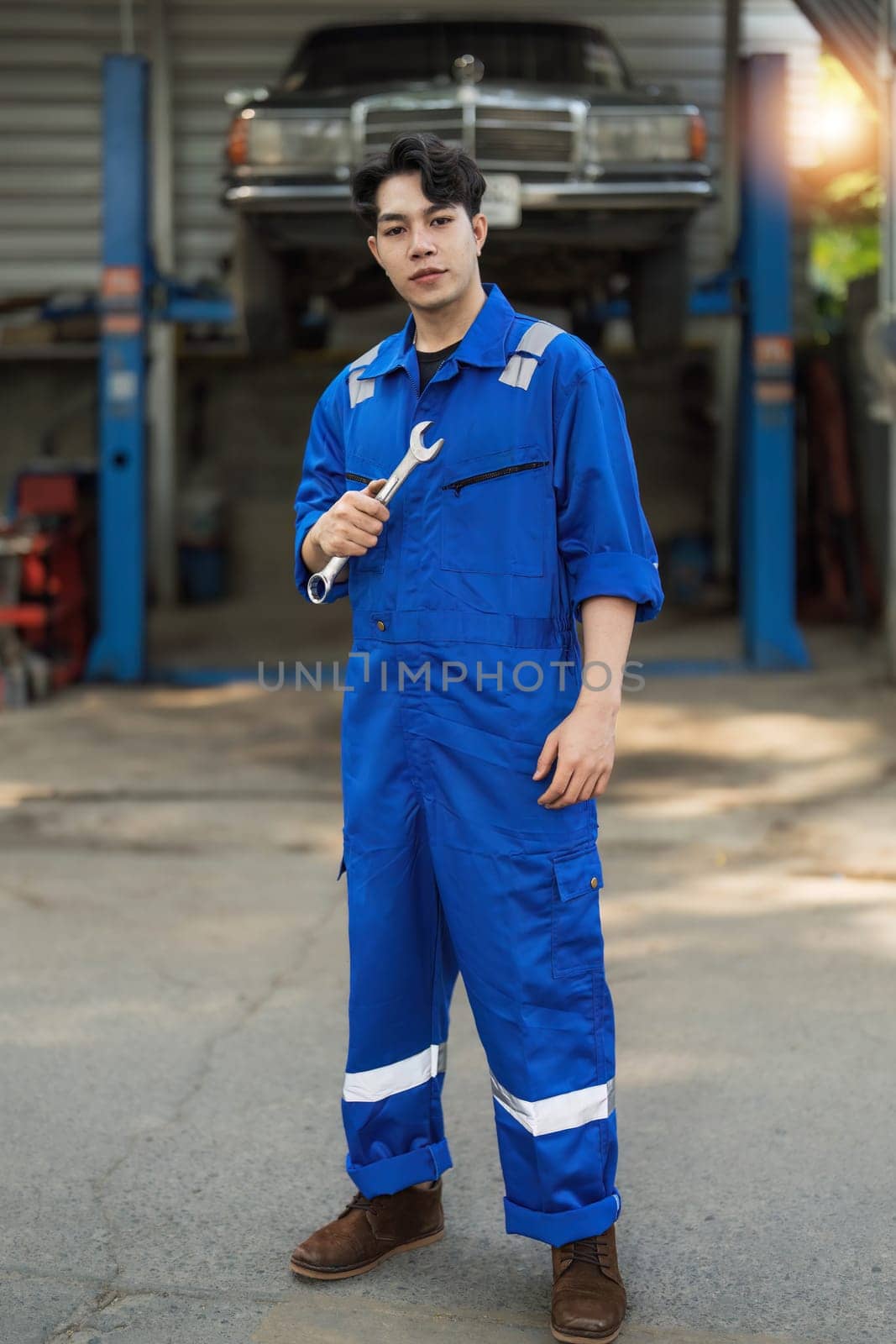 Portrait Shot of a Handsome Mechanic Working on a Vehicle in a Car Service. Professional Repairman is Wearing Gloves and Using a Ratchet Underneath the Car. Modern Clean Workshop...