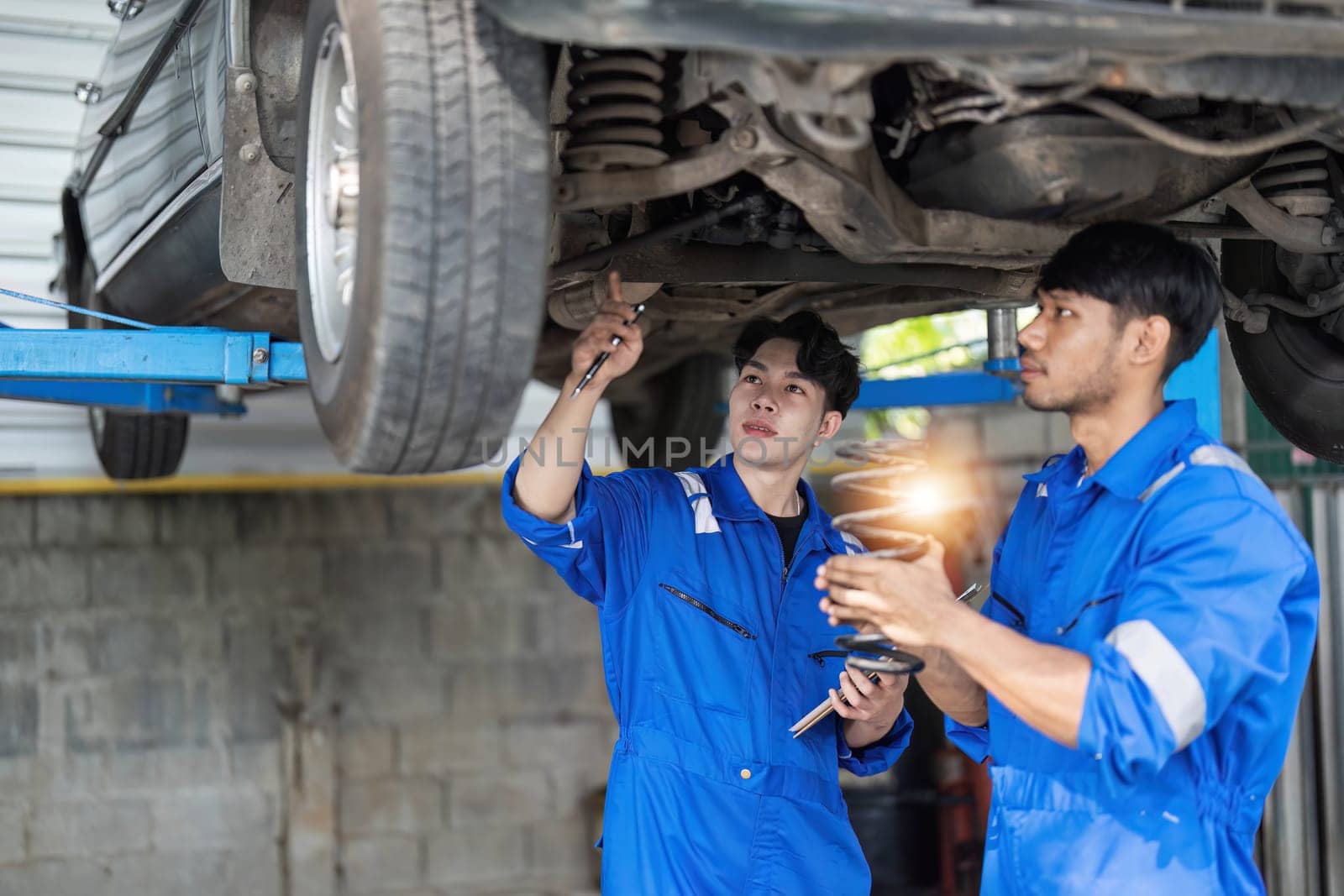 Two Mechanics in a Service are Inspecting a Car After They Got the Diagnostics Results. Female Specialist is Comparing the Data on a Tablet Computer. Repairman is Using a Ratchet to Repair the Faults. by wichayada