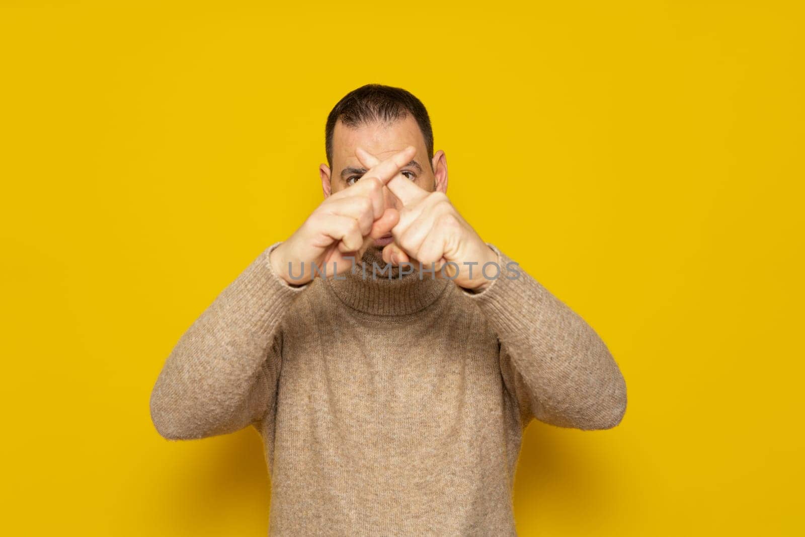 Bearded hispanic man in beige turtleneck shows hands to stop timeout, X fingers. Isolated over yellow background