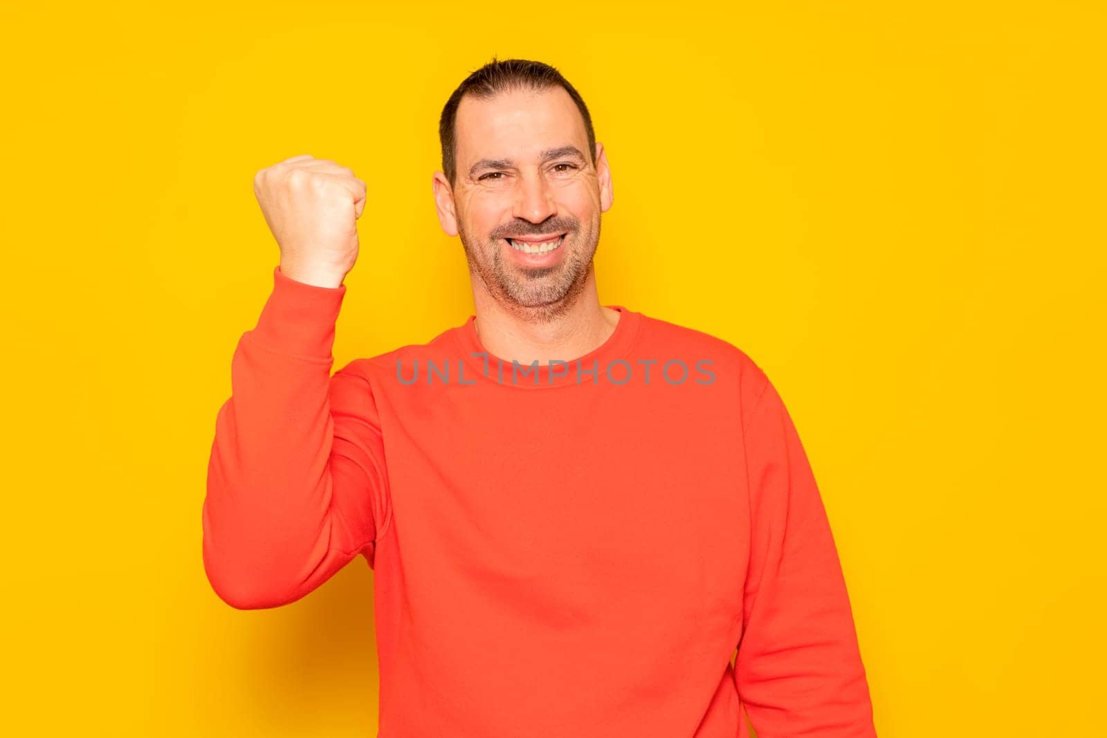 Bearded hispanic man in his 40s wearing a red jumper raising his fist and smiling in victory, isolated over yellow background