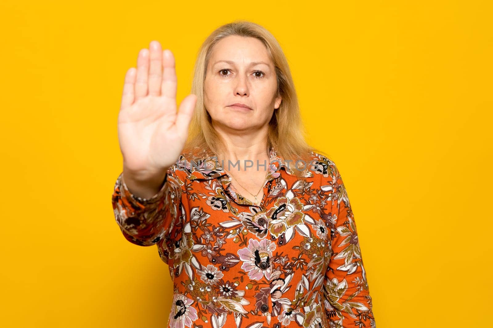 Caucasian blonde woman in a patterned dress raising her palm in a stop sign, isolated over yellow background
