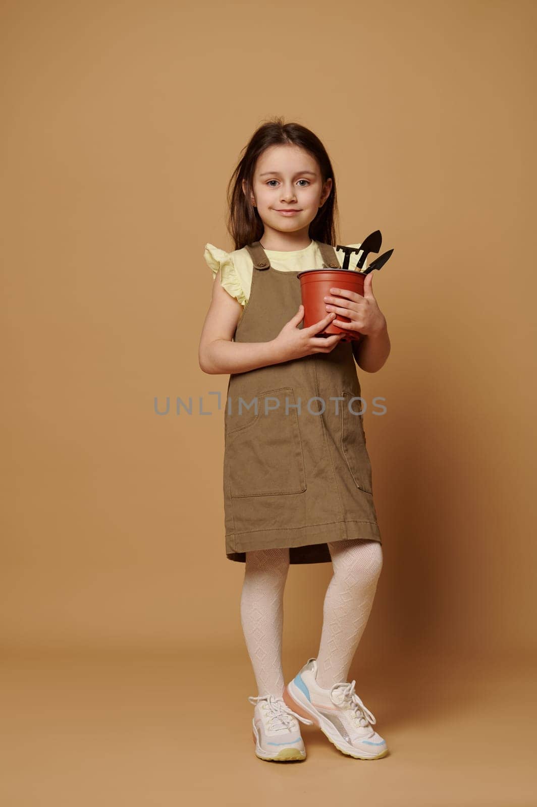 Full-length portrait: lovely little girl in khaki dress, smiles at camera, holding garden shovel and rake in a flower pot isolated over beige studio background. the child dreams of becoming a gardener