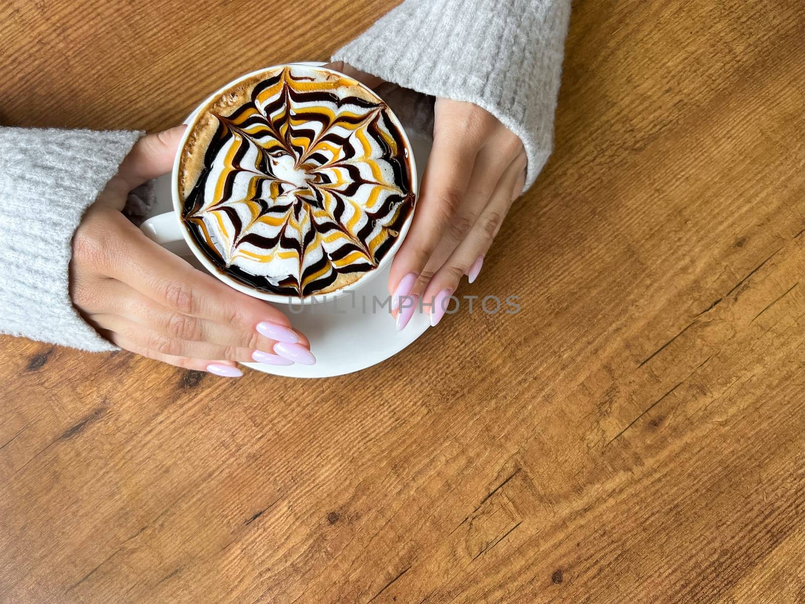 Women's hands hold white cup of coffee with spider web pattern on top of foam, against background of wooden table, close-up, top view