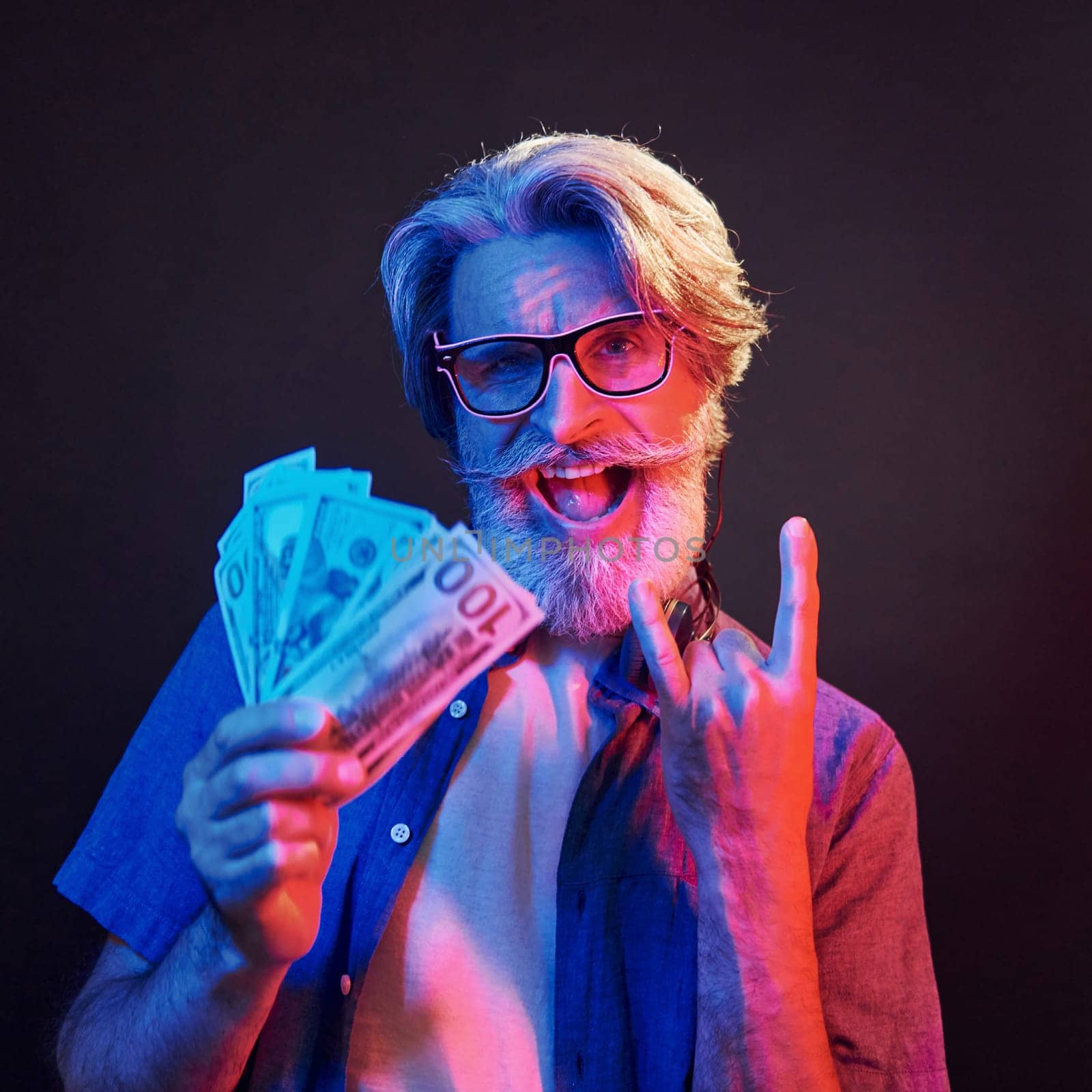 Holding cash. Neon lighting. Stylish modern senior man with gray hair and beard is indoors.