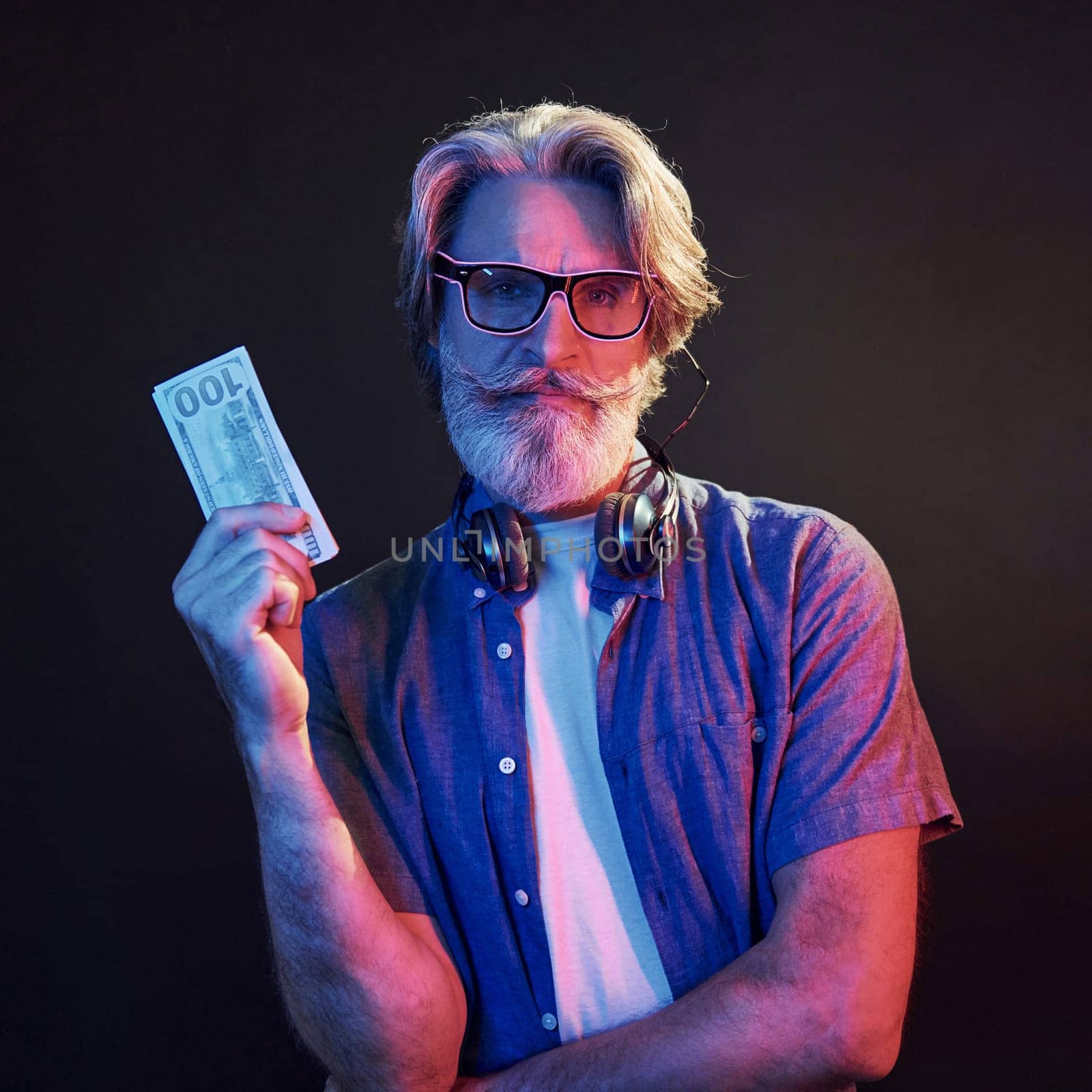 Holding cash. Neon lighting. Stylish modern senior man with gray hair and beard is indoors.