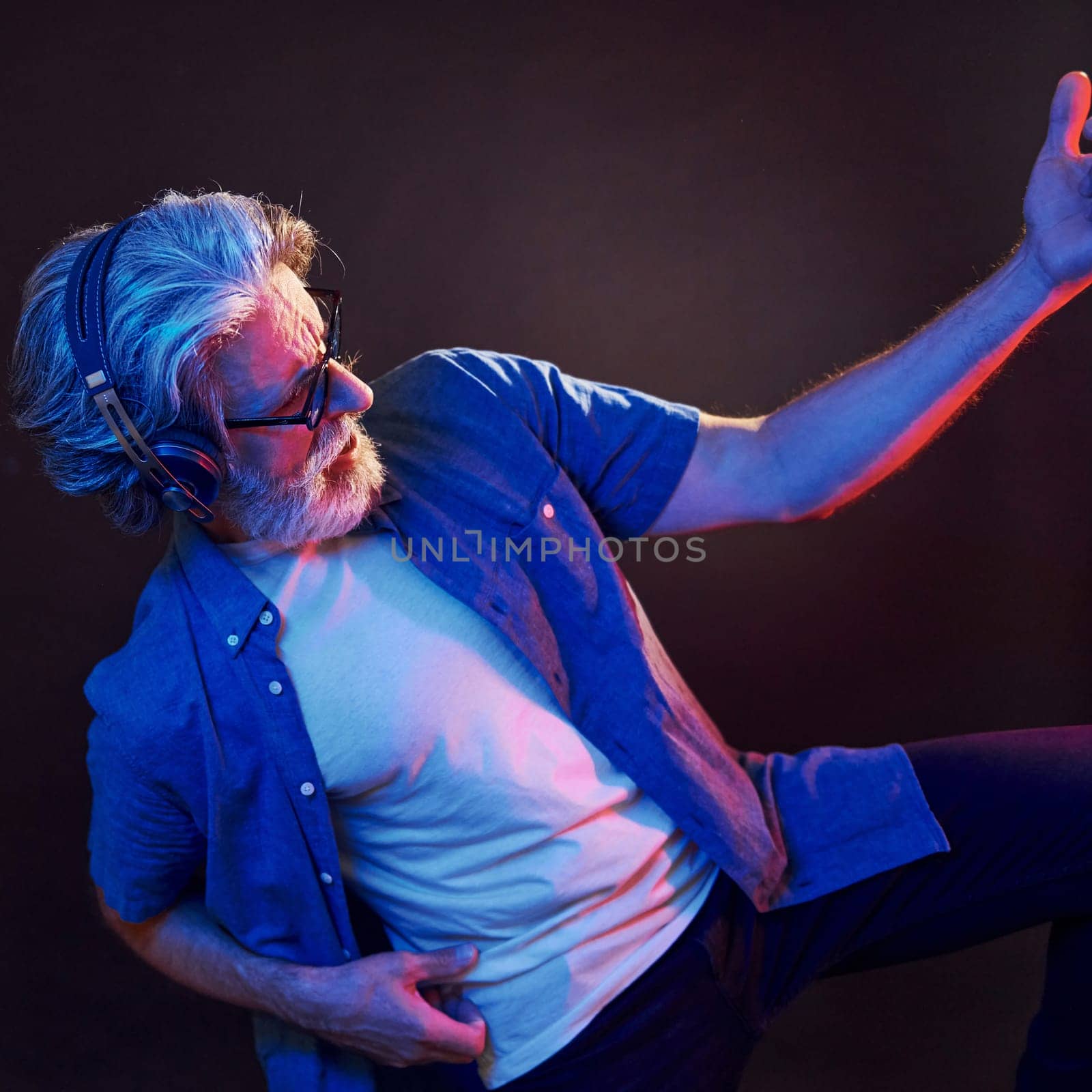 Enjoying music. Neon lighting. Stylish modern senior man with gray hair and beard is indoors.