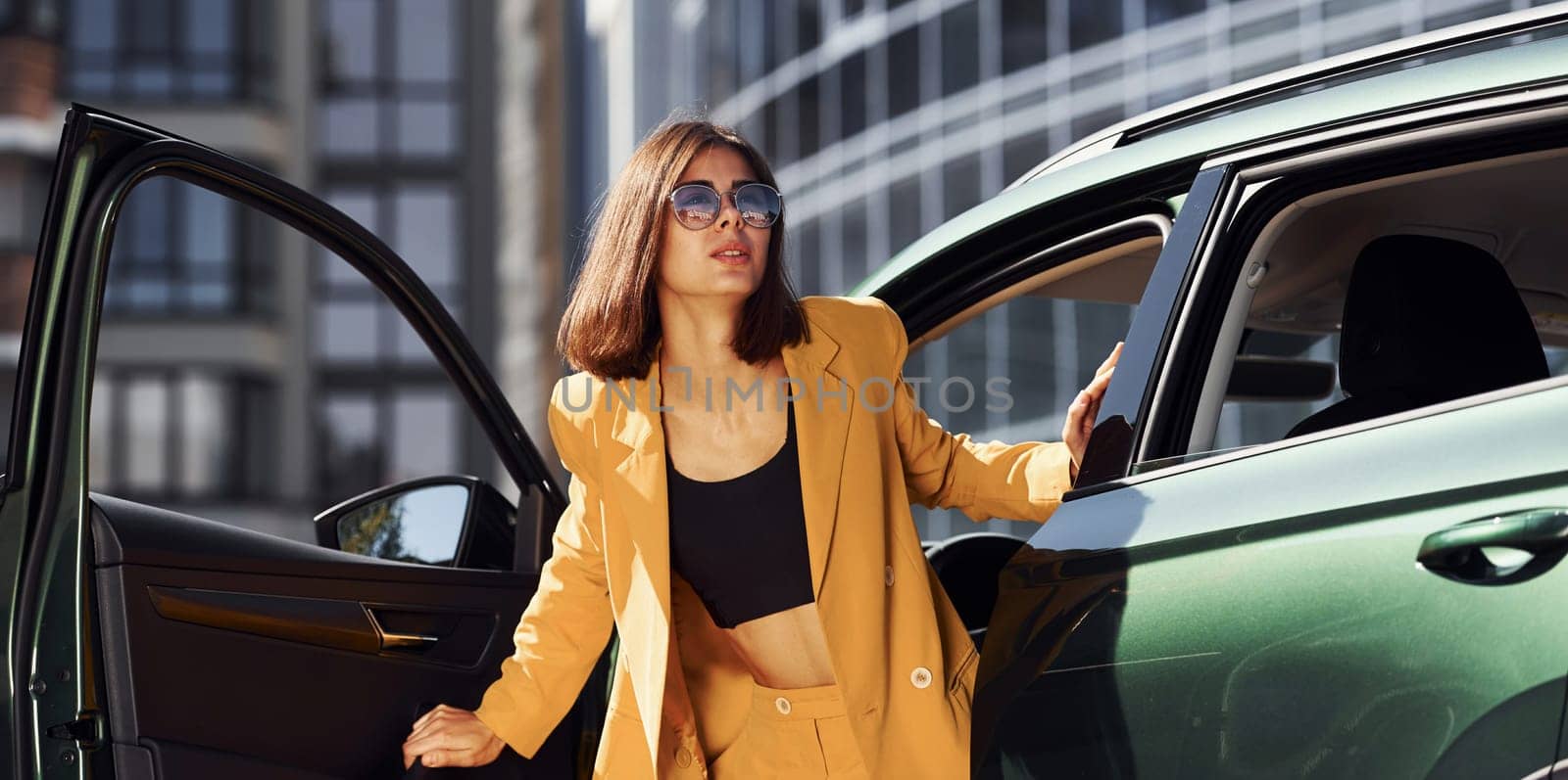 Exits vehicle. Young fashionable woman in burgundy colored coat at daytime with her car.