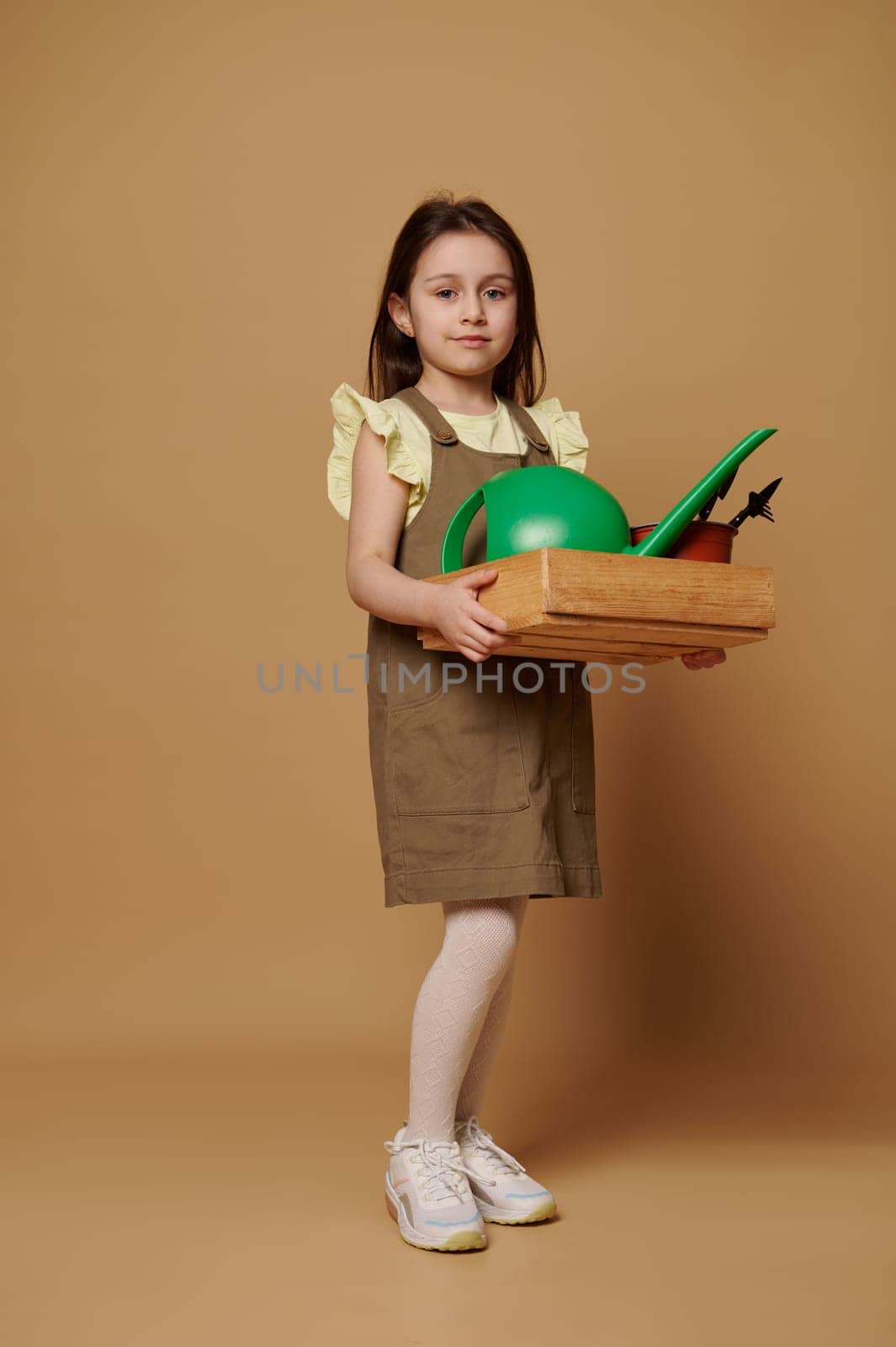 Full-length portrait: lovely child girl enjoys gardening hobby, smiling at camera, carrying a wooden crate with garden tools and watering can, ready for sowing and planting, isolated beige background