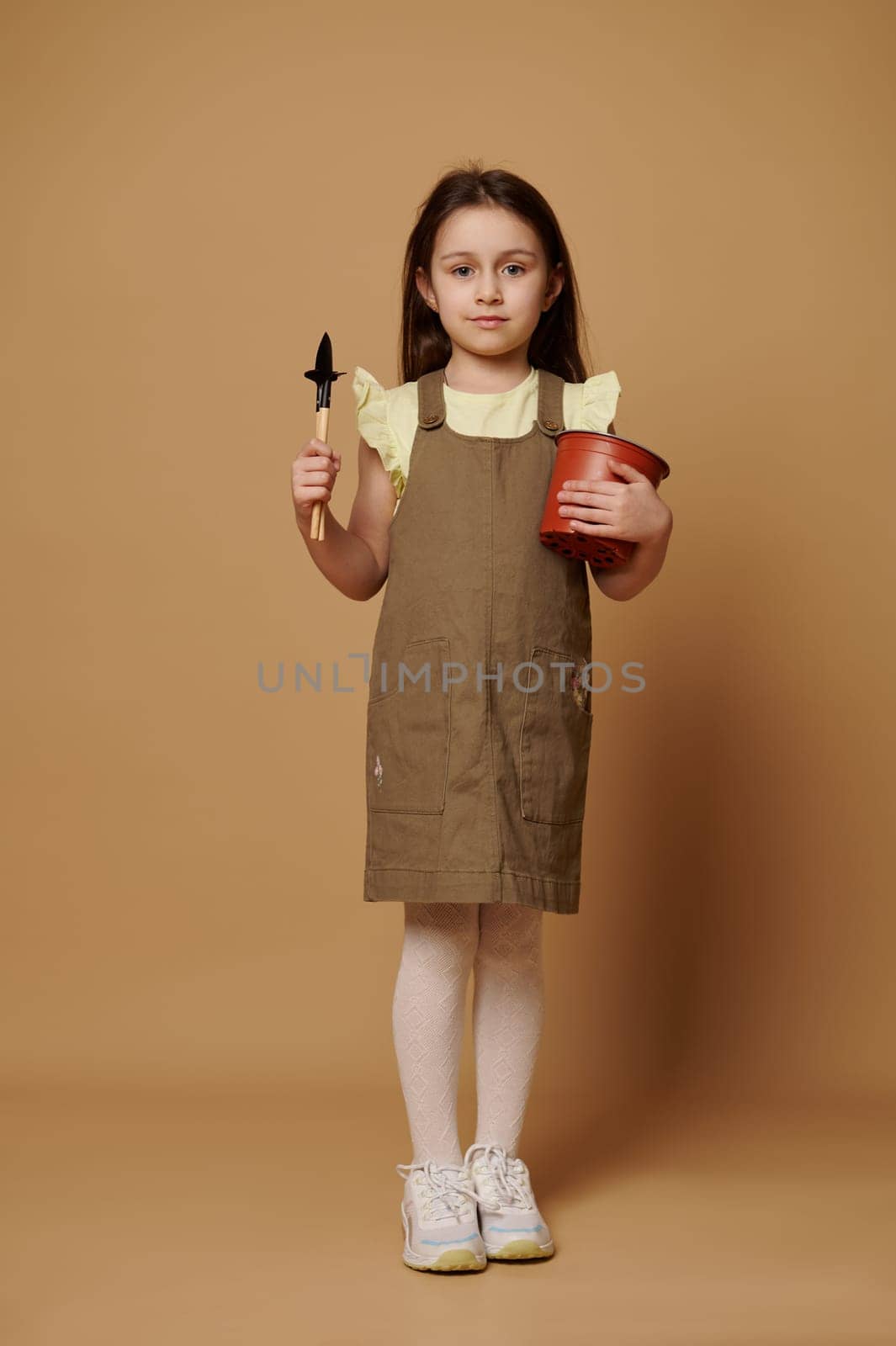 Caucasian happy little child girl smiling broadly, looking at camera, holding a flower pot, garden shovel and rake, isolated over beige background. Full-length of a cute kid dreaming becoming gardener