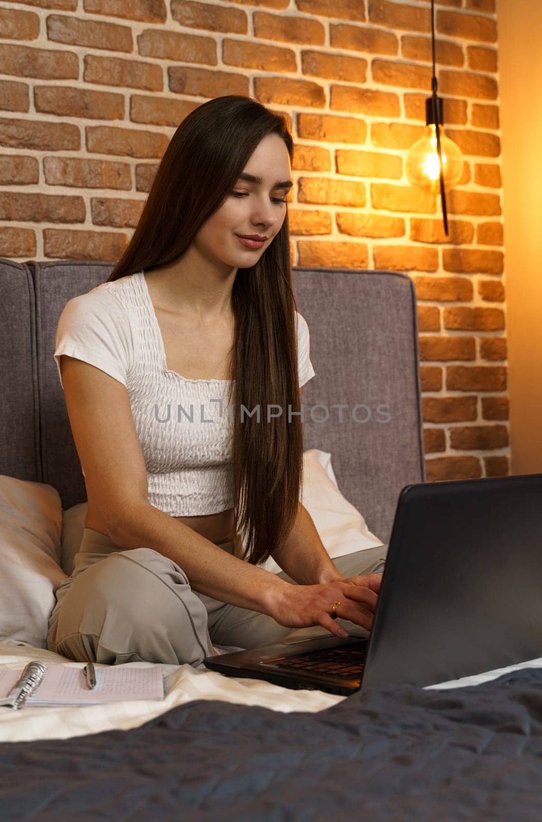 Happy beautiful woman is sitting on the bed and working on the laptop. Vertical frame.