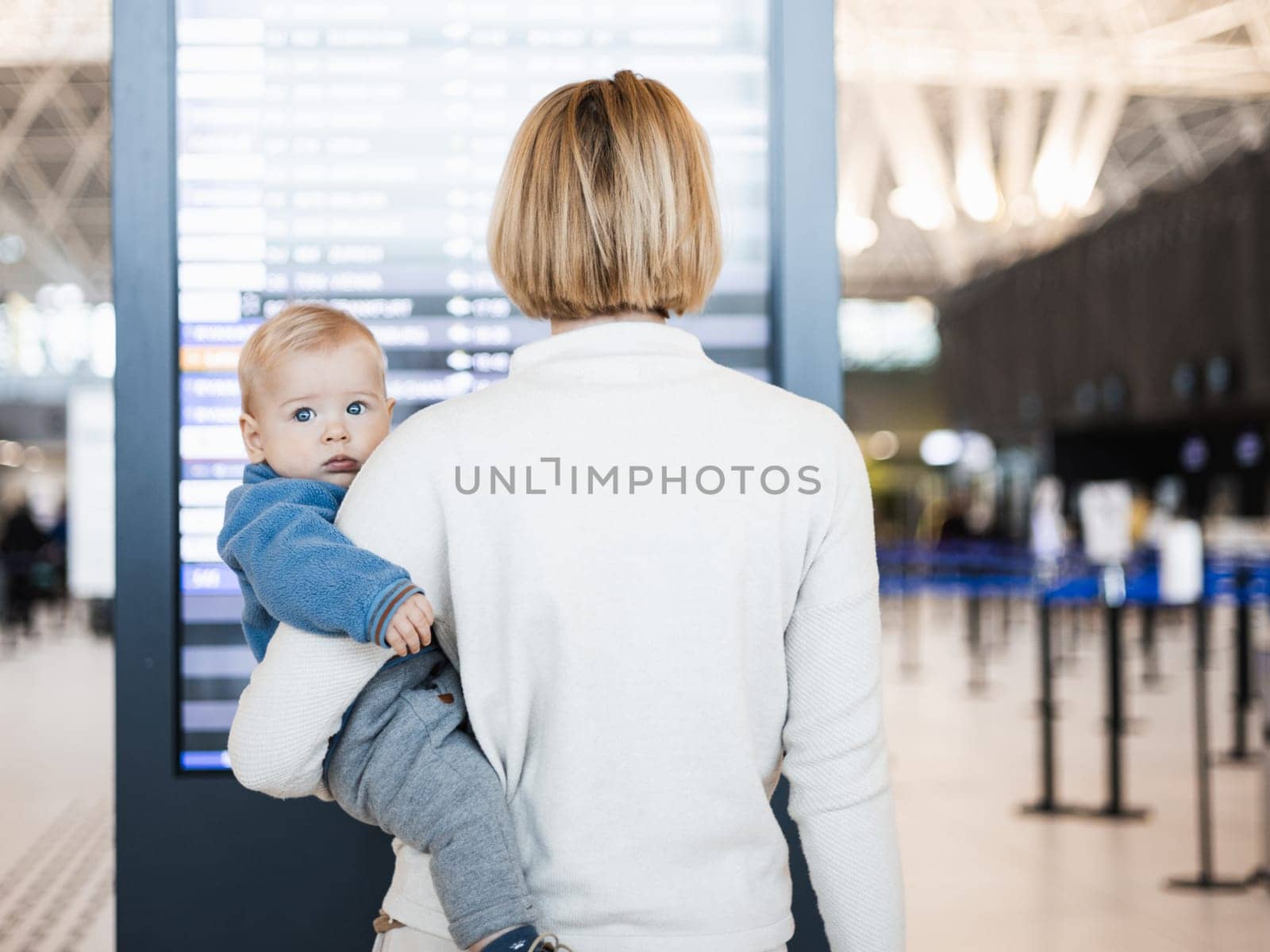 Mother traveling with child, holding his infant baby boy at airport terminal, checking flight schedule, waiting to board a plane. Travel with kids concept. by kasto