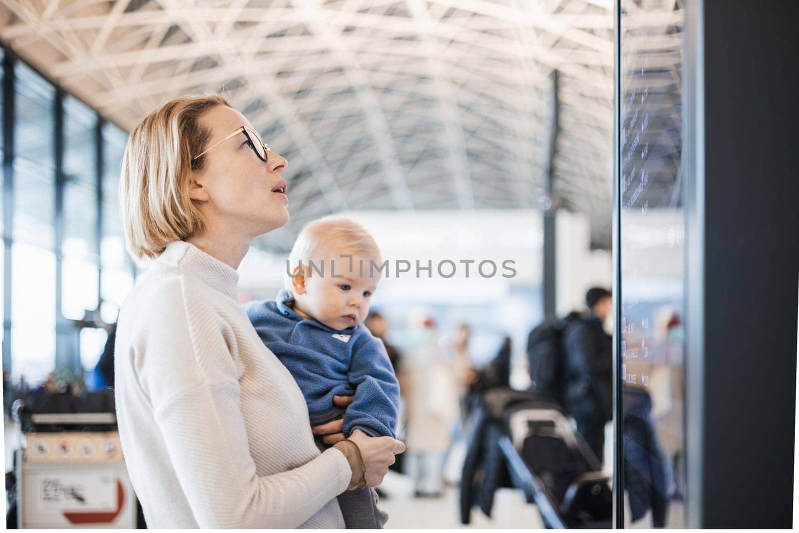 Mother traveling with child, holding his infant baby boy at airport terminal, checking flight schedule, waiting to board a plane. Travel with kids concept