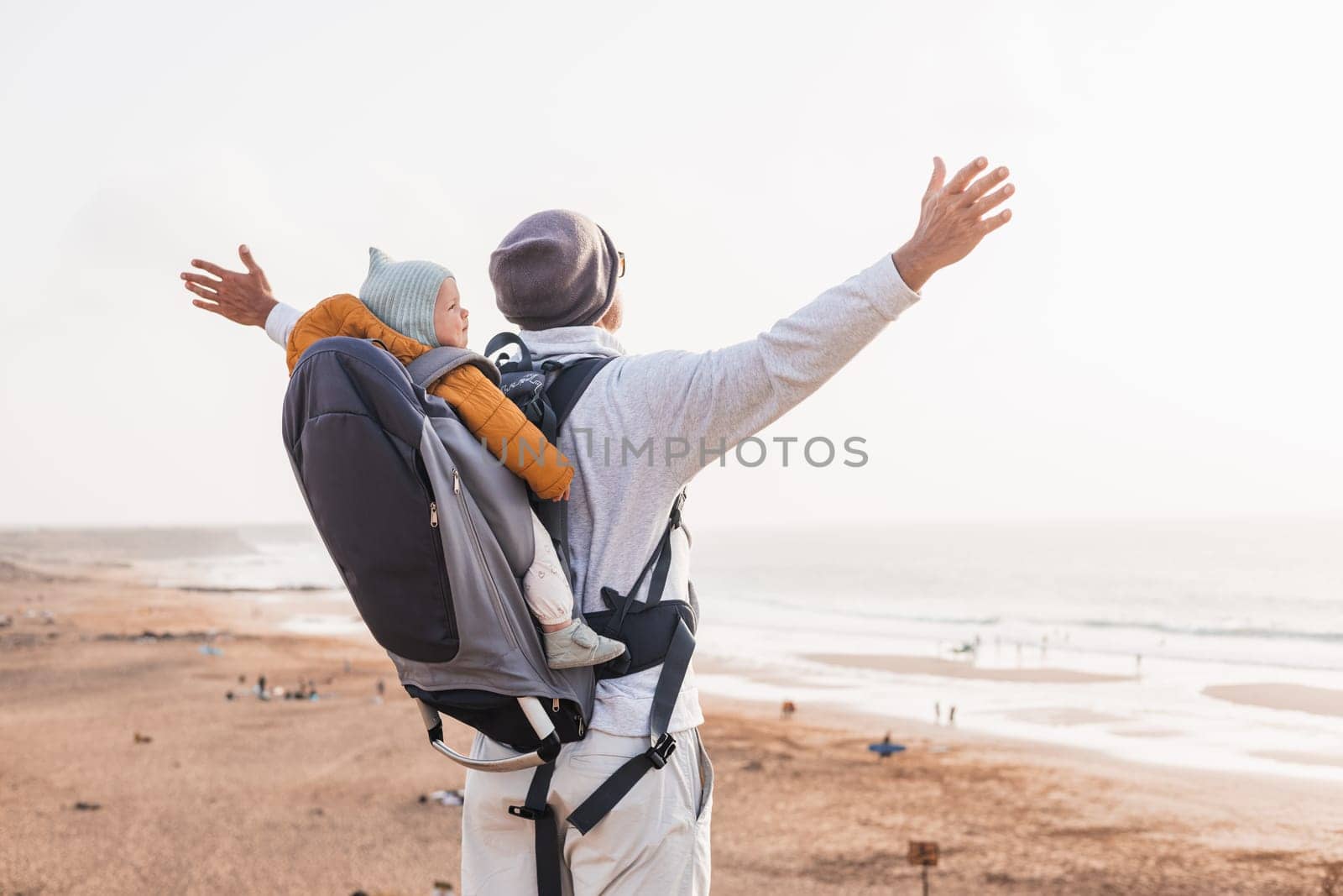 Young father rising hands to the sky while enjoying pure nature carrying his infant baby boy son in backpack on windy sandy beach. Family travel concept. by kasto