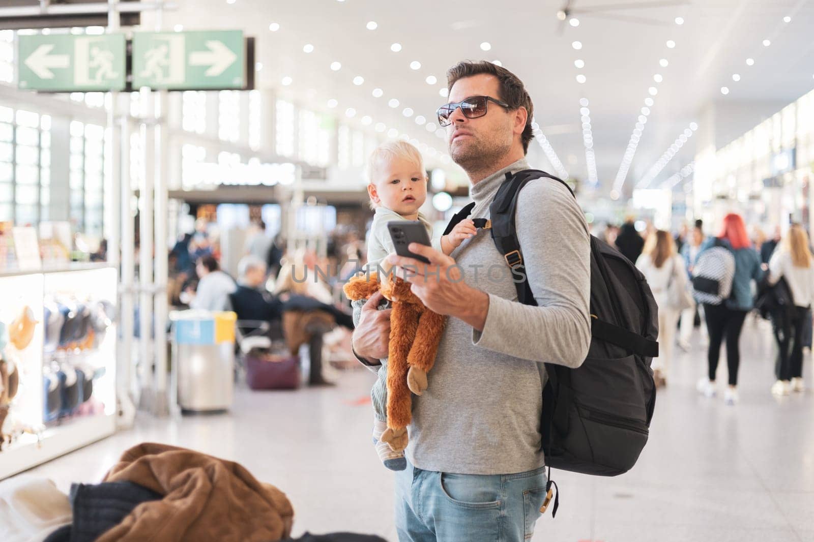 Father traveling with child, holding his infant baby boy at airport terminal, checking flight schedule, waiting to board a plane. Travel with kids concept