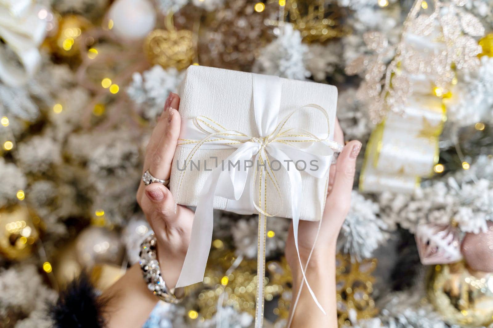 Female hands hold a gift box tied with a ribbon on the background of a Christmas tree, the concept of christmas and gifts. by Matiunina