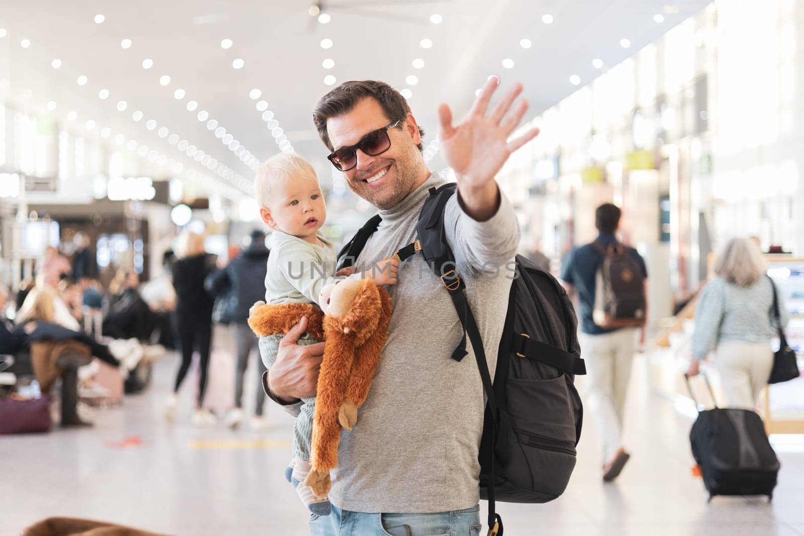 Father traveling with child, holding his infant baby boy at airport terminal waiting to board a plane waving goodby. Travel with kids concept