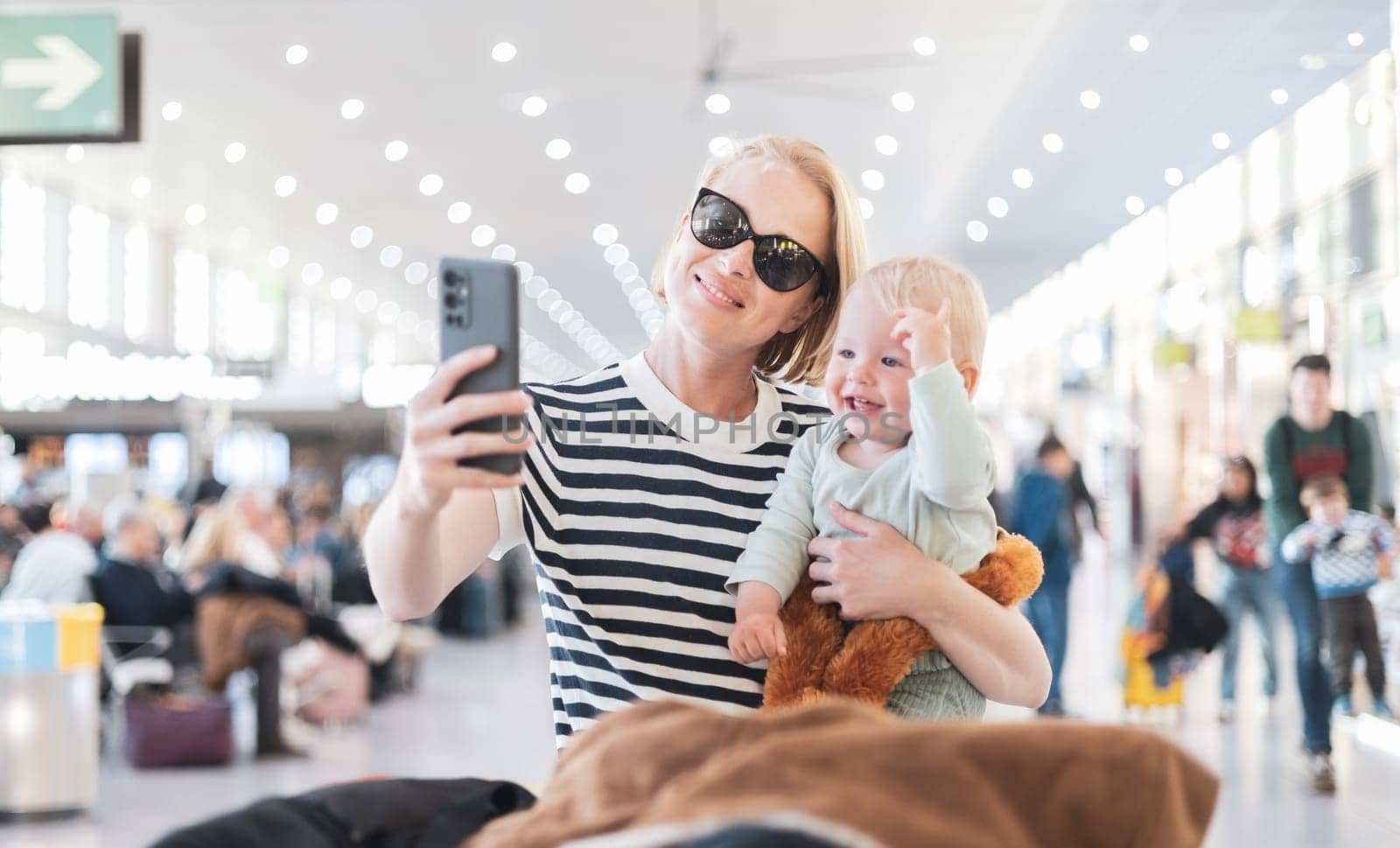Mother taking selfie with mobile phone, while traveling with child, holding his infant baby boy at airport, waiting to board a plane. Travel with kids concept