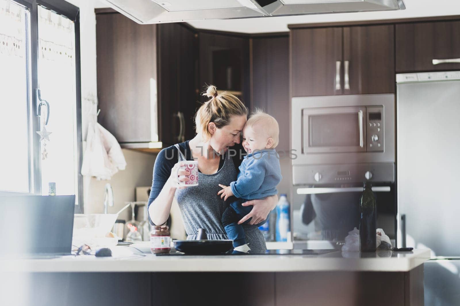 Happy mother holding her little infant baby boy while drinking morning coffee and making pancakes for breakfast in domestic kitchen. Family lifestyle, domestic life concept