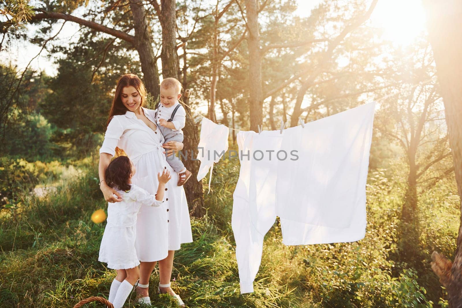 White clothes hanging on the rope to dry. Young mother with her little daughter and son is outdoors in the forest. Beautiful sunshine.
