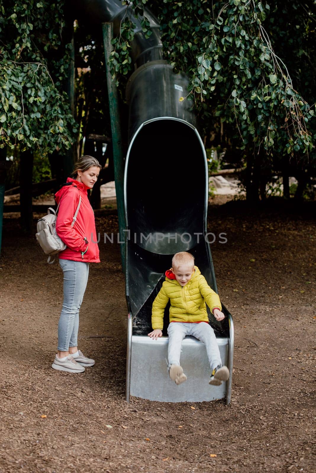 Curious toddler boy in warm clothes playing on the playground slide with his mommy. Caucasian mother stand and take care little boy play slide in fun park of garden and they look happy to have enjoy time together. Pretty mum catching male kid, riding on slide at modern courtyard of city residential high-rise buildings. Mother catching little son while standing near slide on playground. Concept of parenthood