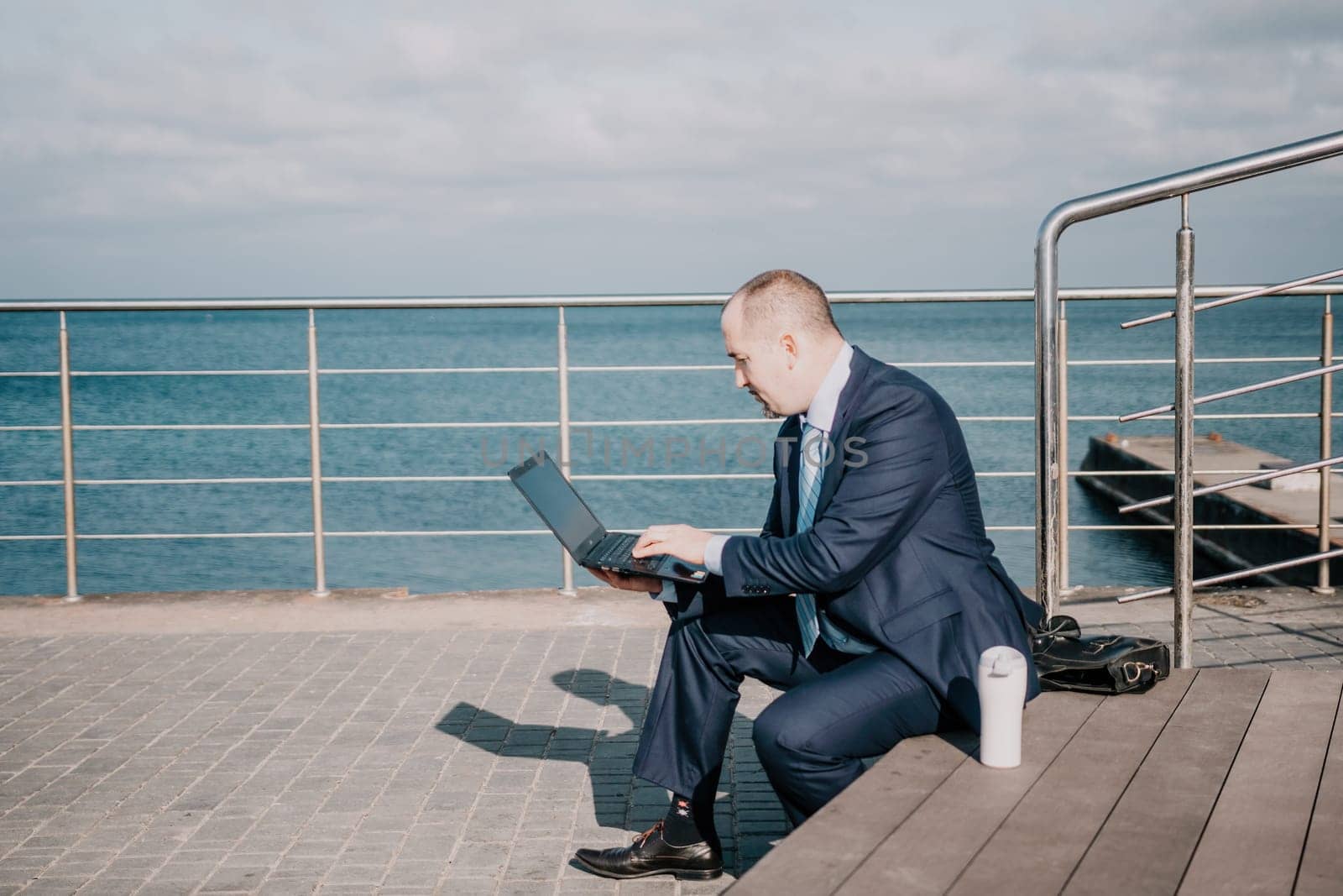 Confident middle age businessman working remotely online, typing on a laptop keyboard while sitting on a beach at sunset. Working remotely on vacation, running an online business from a distance. by panophotograph