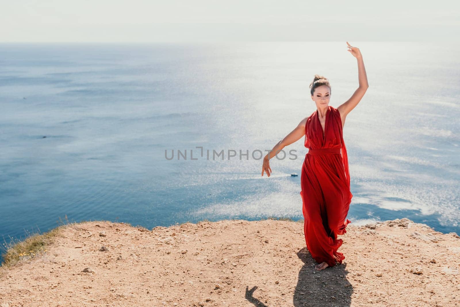 Side view a Young beautiful sensual woman in a red long dress posing on a rock high above the sea during sunrise. Girl on the nature on blue sky background. Fashion photo.