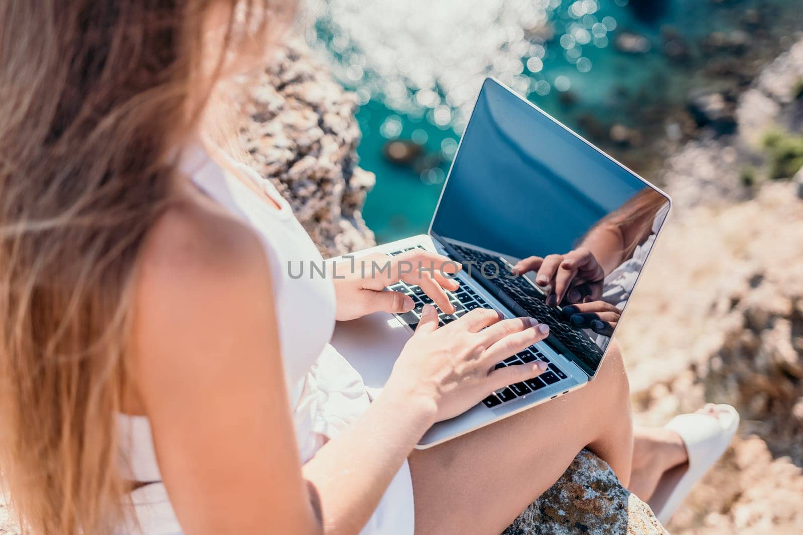 Successful business woman in yellow hat working on laptop by the sea. Pretty lady typing on computer at summer day outdoors. Freelance, travel and holidays concept.