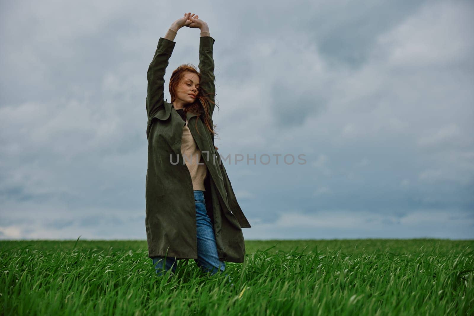 a woman in a long coat stands in a field against a cloudy sky with her back to the wind with her hands raised behind her head by Vichizh