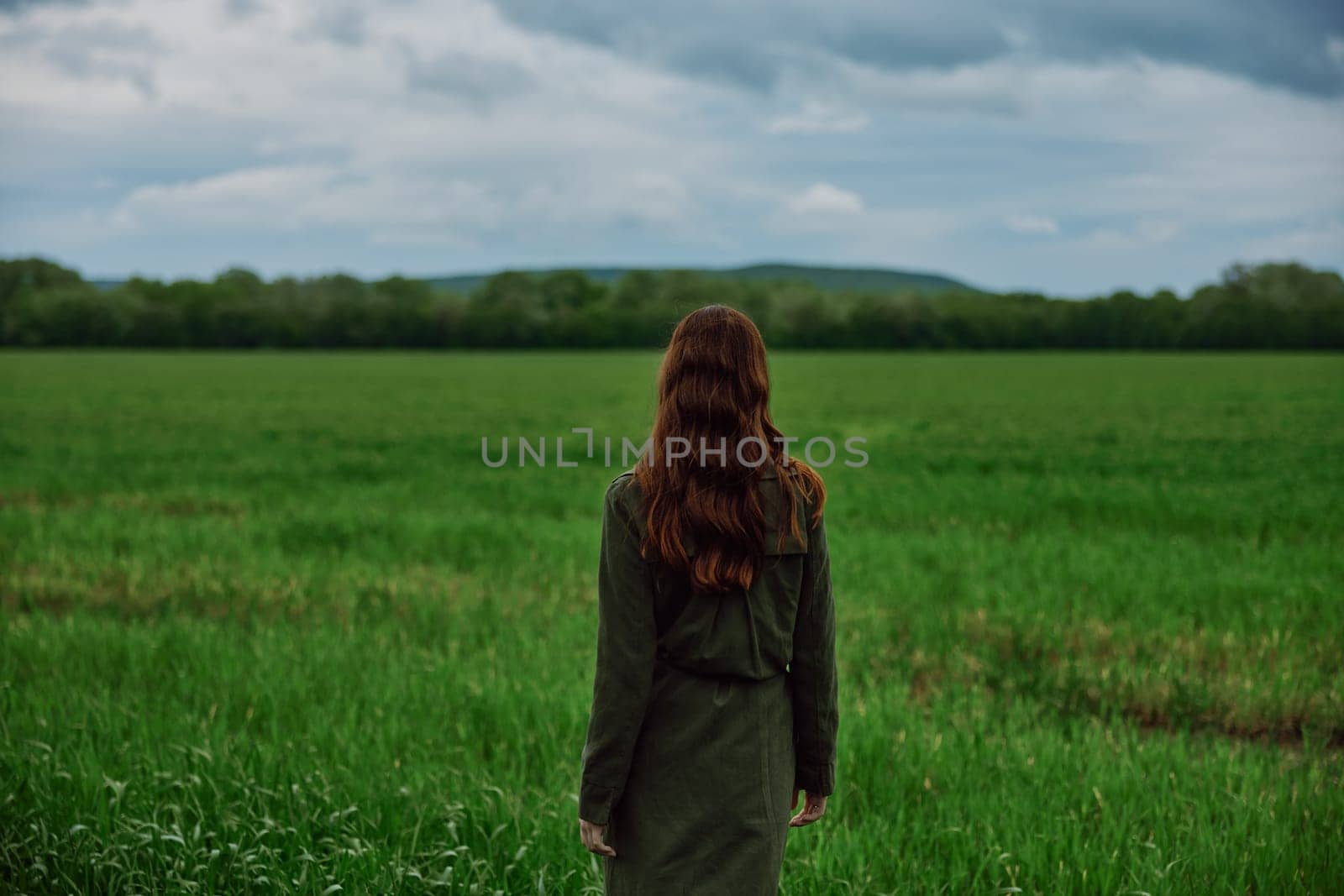 a woman with beautiful, long, red hair stands with her back to the camera in a green field in rainy, spring weather in a long raincoat by Vichizh
