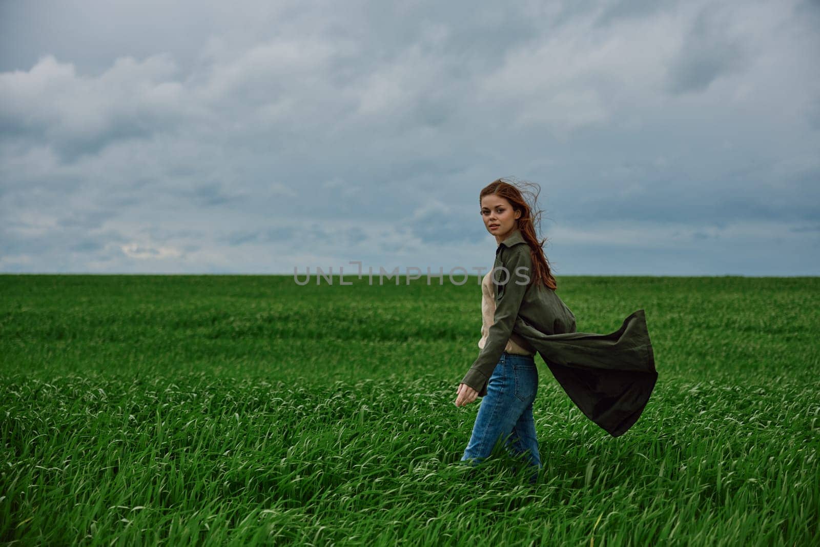 A woman stands in a green field in a raincoat and looks at the camera. Strong wind, flying hair, freedom and harmony with nature. by Vichizh