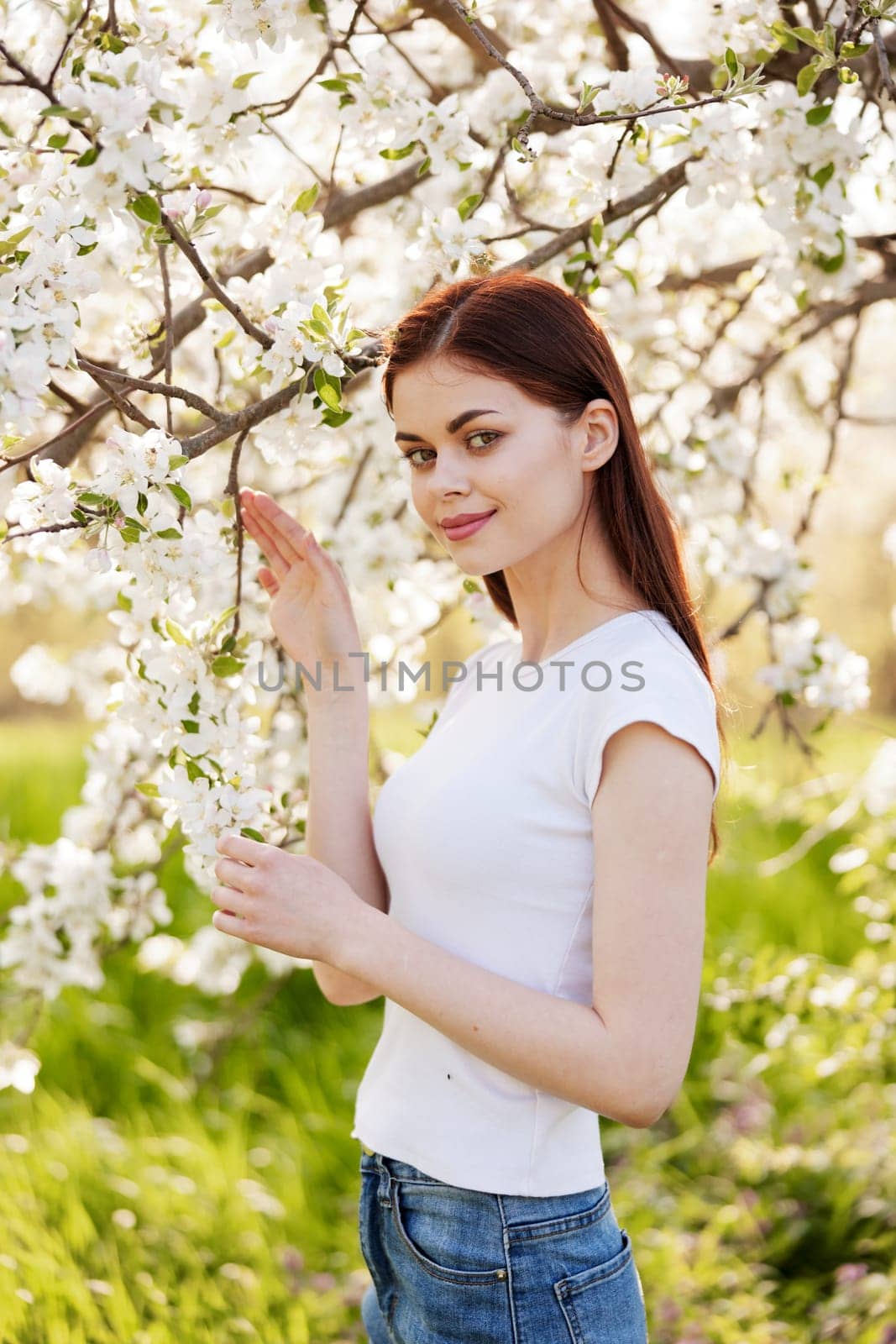 portrait of a happy woman with red hair in casual clothes touching flowers on the branches of a fruit tree by Vichizh