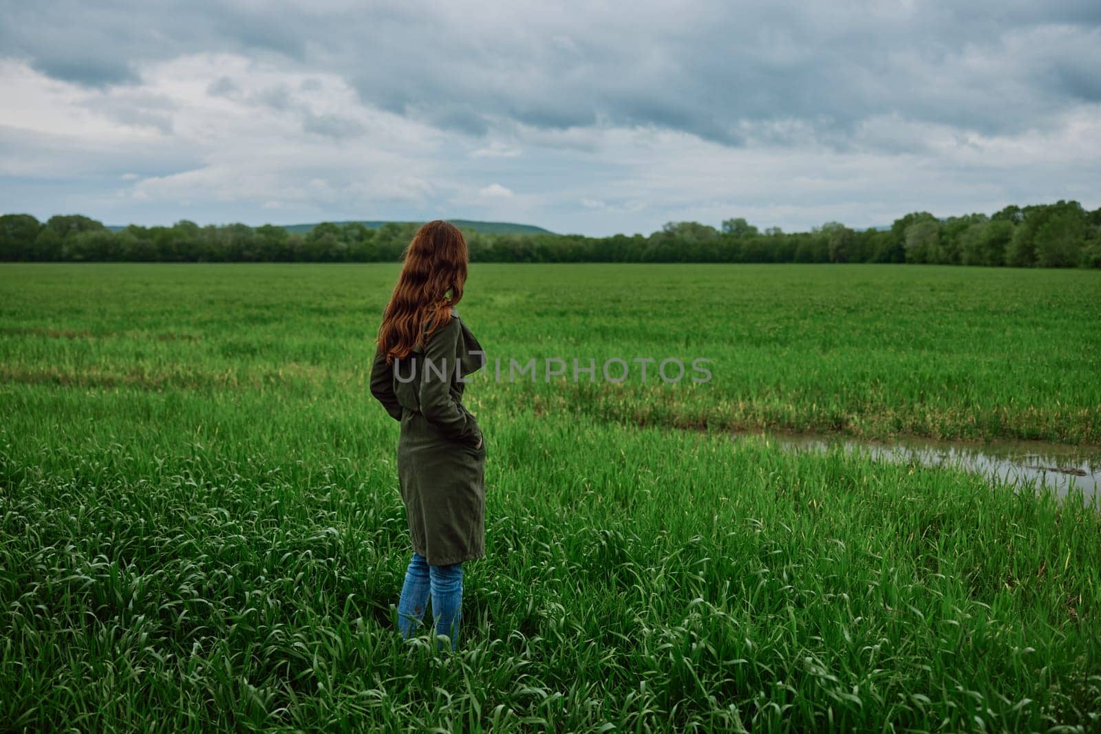 a woman with beautiful, long, red hair stands with her back to the camera in a green field in rainy, spring weather in a long raincoat by Vichizh