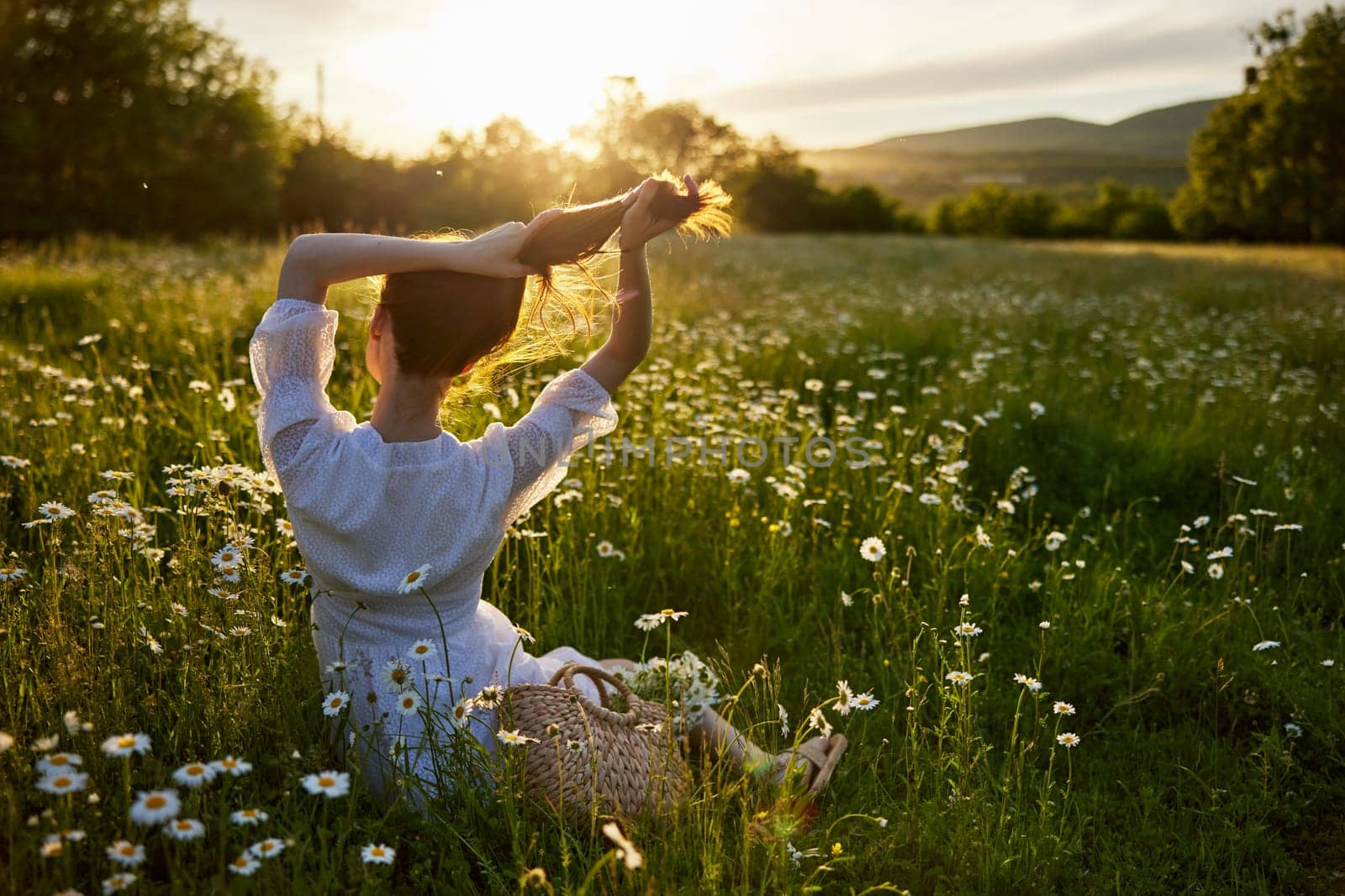 a red-haired woman in a light dress sits with her back to the camera in a field of daisies straightening her hair with her hands against the backdrop of the setting sun by Vichizh