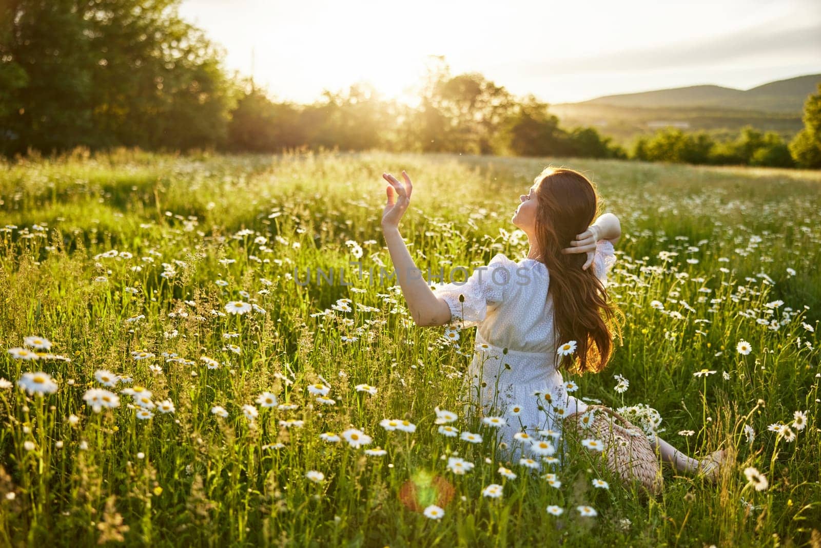 a red-haired woman in a light dress sits with her back to the camera in a field of daisies straightening her hair with her hands against the backdrop of the setting sun by Vichizh