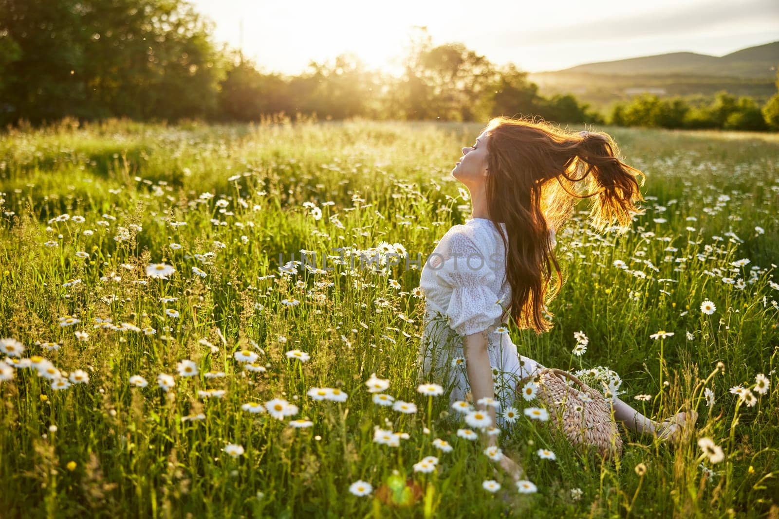 a red-haired woman in a light dress sits with her back to the camera in a field of daisies straightening her hair with her hands against the backdrop of the setting sun. High quality photo