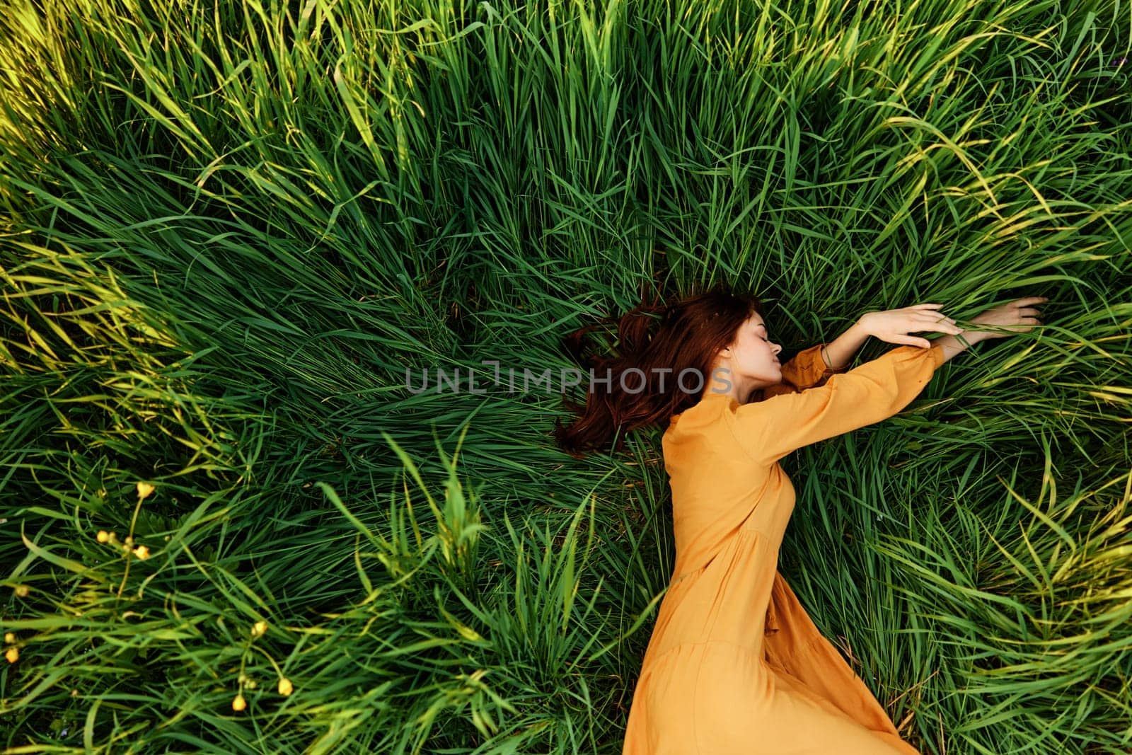 an elegant woman lies in the tall green grass in a long summer orange dress and relaxed posing with her hands enjoying nature and sunny weather with her eyes closed. High quality photo