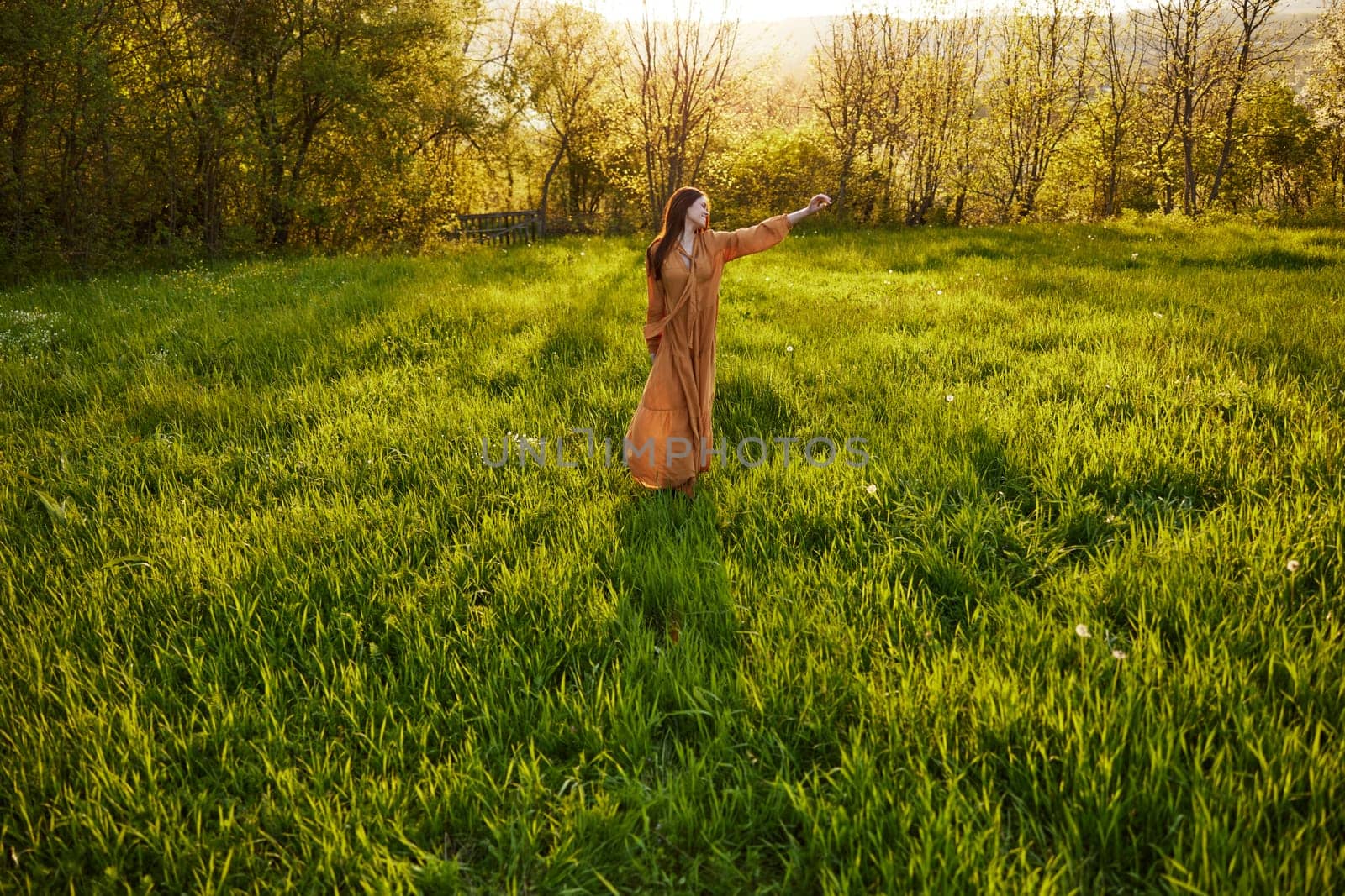 an attractive, slender, red-haired woman stands far away in a wide, green field during sunset in a long orange dress enjoying unity with nature and relaxation, raising her arms to the sides by Vichizh