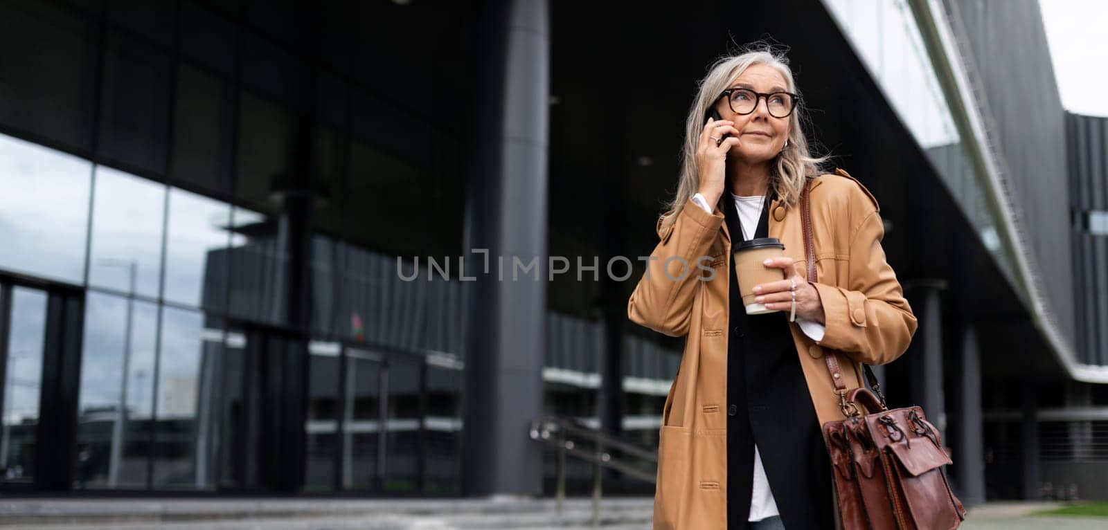 photo of a mature adult female economist with gray hair in a brown coat during a break against the backdrop of a business center.