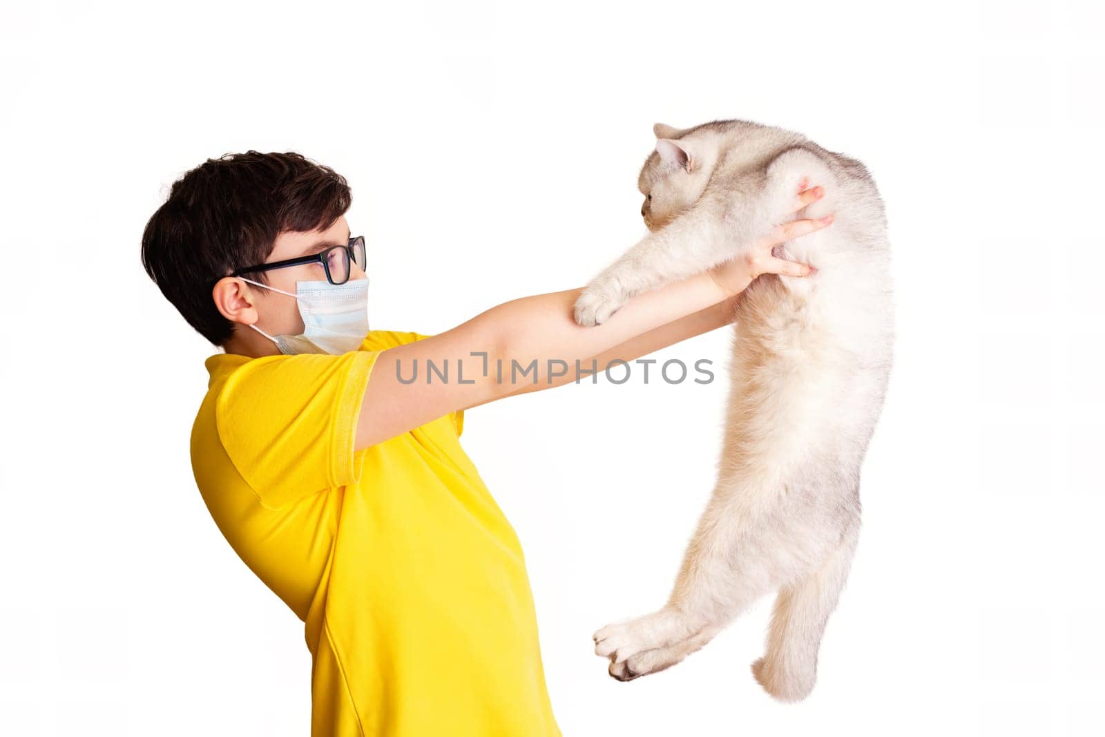 A brunette boy in a medical mask, in yellow t-shirt, holds his white cat at arms length, isolated on white background. Allergy to animal fur concept. Close up. Copy space