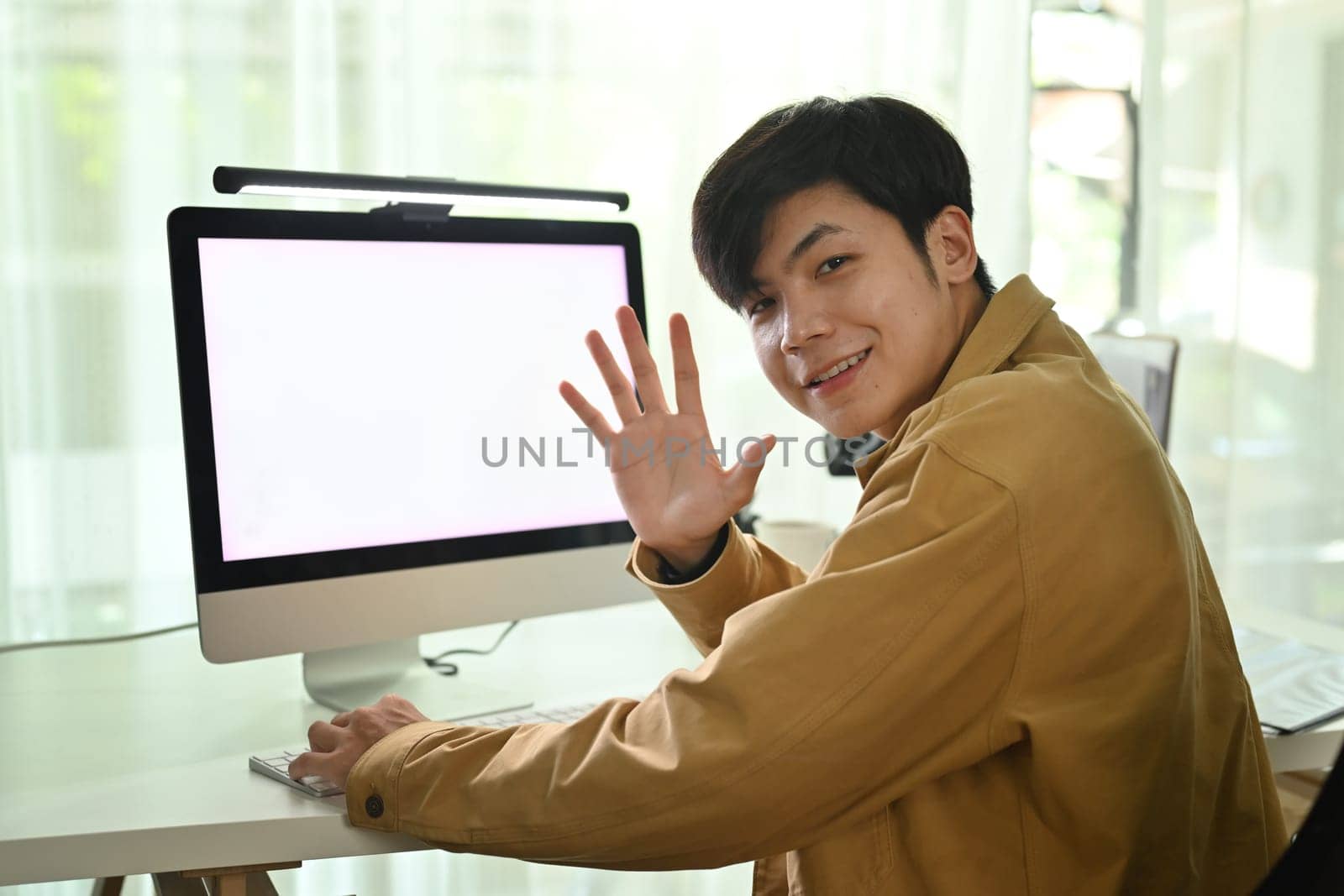 Cheerful asian man worker waving hand at camera, greeting someone while sitting in modern office.