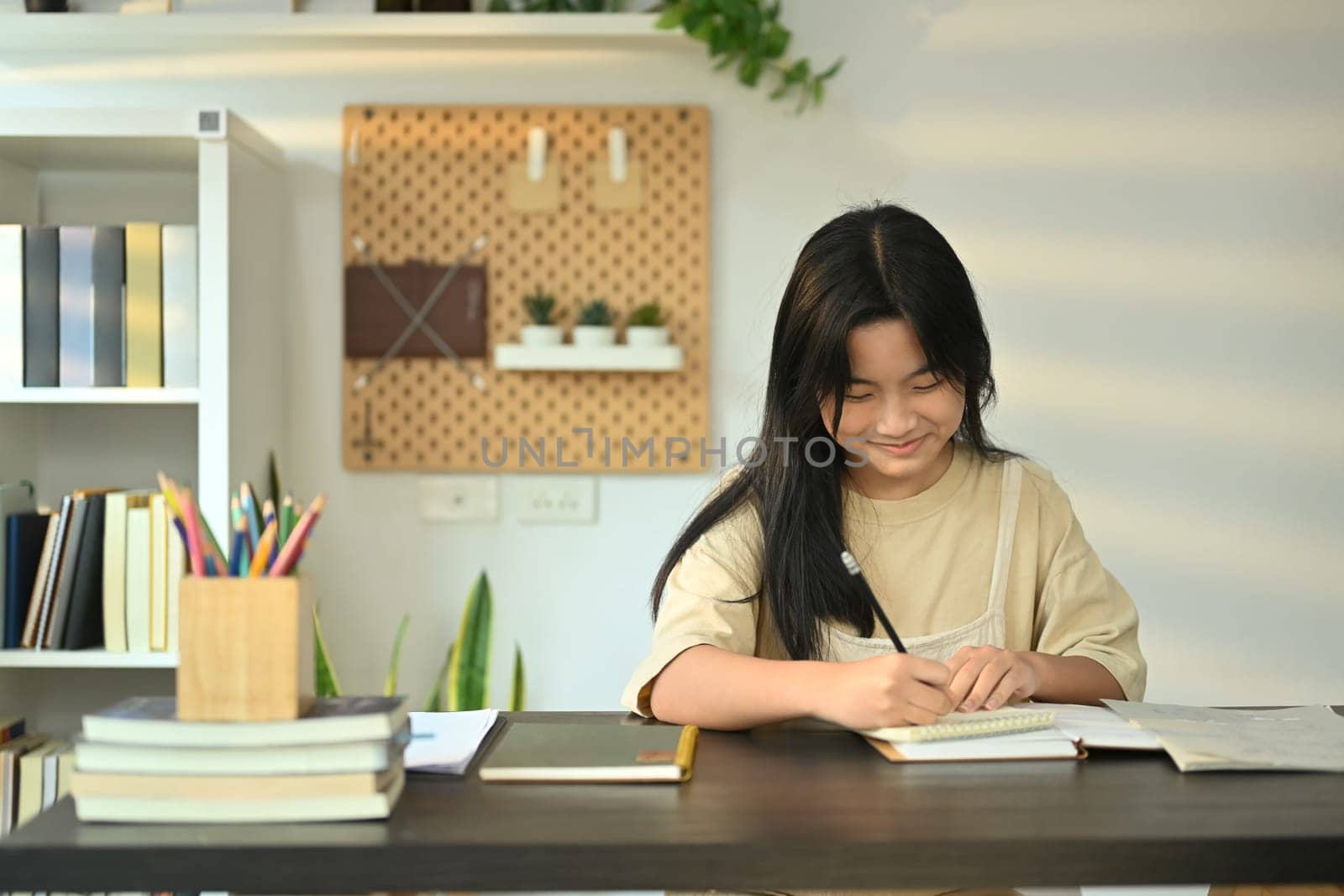 Focused teenage girl doing homework at home, writing down in notebook. Learning and education concept by prathanchorruangsak