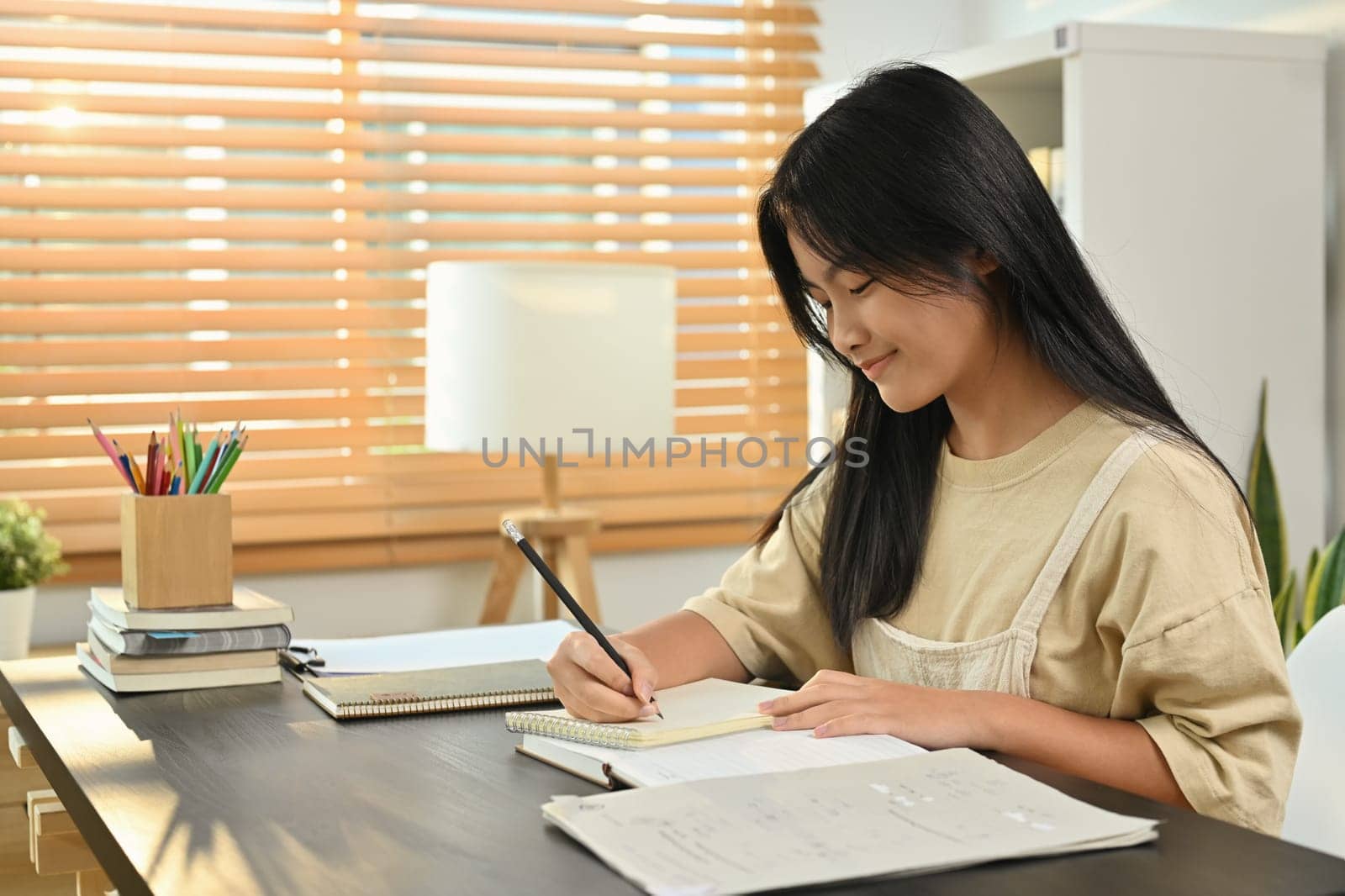 Cute asian teenage student, doing homework, writing task, taking notes on desk at home by prathanchorruangsak