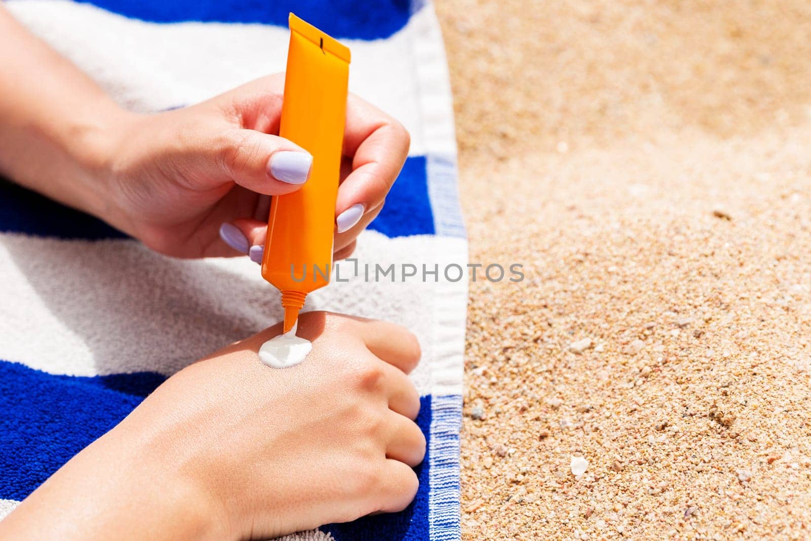 Attractive woman is lying on a striped towel at the beach and squeezing sunblock from a tube on her hand.