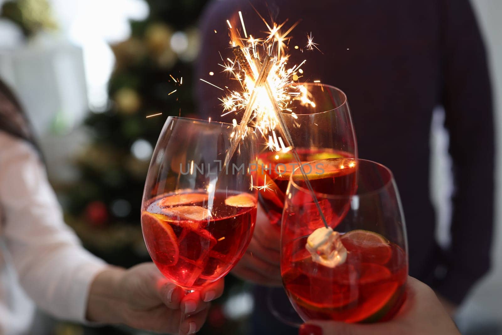 People hold glasses with cocktails with sparklers in front of Christmas tree closeup by kuprevich