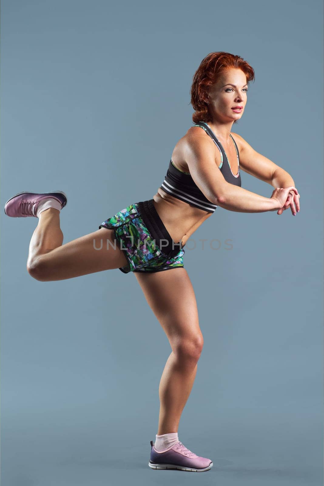 Mature athletic redhead woman in sportswear t-shirt and shorts doing fitness exercises, gray studio background